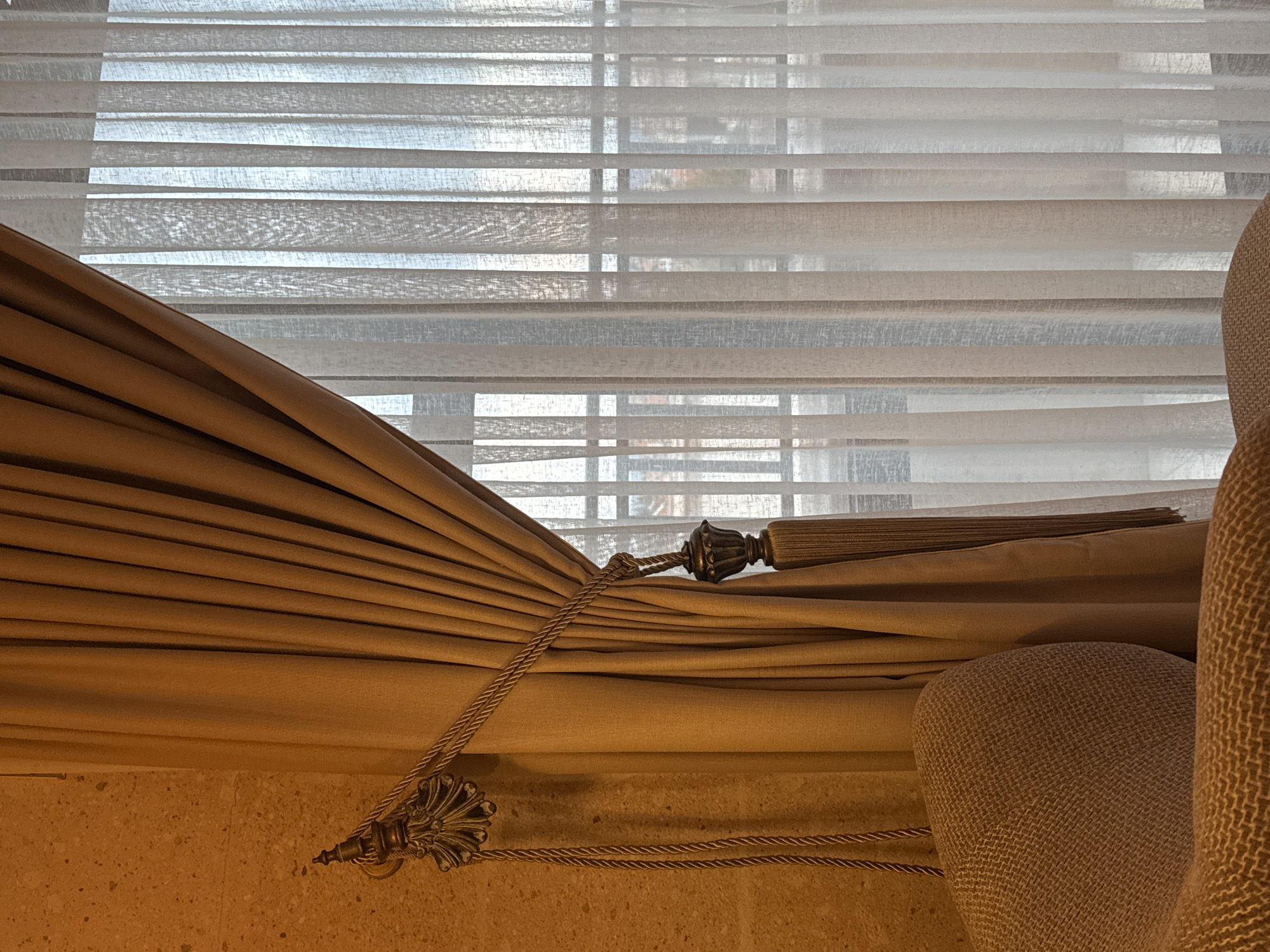 A close-up, indoor shot shows a window with horizontal blinds partially covered by a draped, light-brown curtain. The curtain is held back with a decorative rope and tassel. A chair, upholstered in a similar light-brown fabric with a textured pattern, is partially visible on the right side. The wall, visible at the bottom left, has a rough, textured surface in a beige color. The scene appears to be in a residential setting. The location, Ürgüp, Turkey, is not discernible within the image itself, though it is included within the instructions.