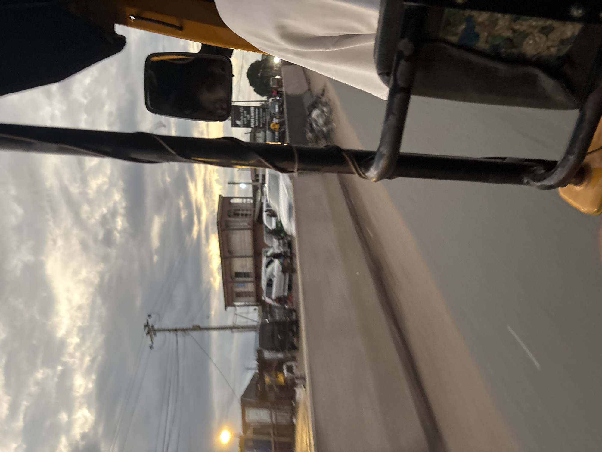 The image captures a view from the passenger seat of a yellow auto-rickshaw, likely in Onitsha, Nigeria, as indicated by the context. The sky above is filled with dramatic, overcast clouds, illuminated by the setting sun, casting a warm glow on the scene. A power pole with wires is visible in the distance, a common sight in urban Nigerian landscapes.
The road ahead is lined with a concrete barrier, beyond which a row of buildings and various vehicles are present. These include a light-colored minivan, several other cars, and what appears to be a dark colored van. Some of these vehicles seem to be stationary, possibly in traffic or parked. Along the roadside, there are some makeshift structures and piles of debris.
The auto-rickshaw itself features a black metal frame that partially obstructs the view. A side mirror is attached to the frame. The passenger's white shirt sleeve is visible in the foreground, suggesting the perspective of someone riding within the vehicle. The overall atmosphere suggests a busy urban environment at dusk, with the characteristic streetscape of Onitsha.