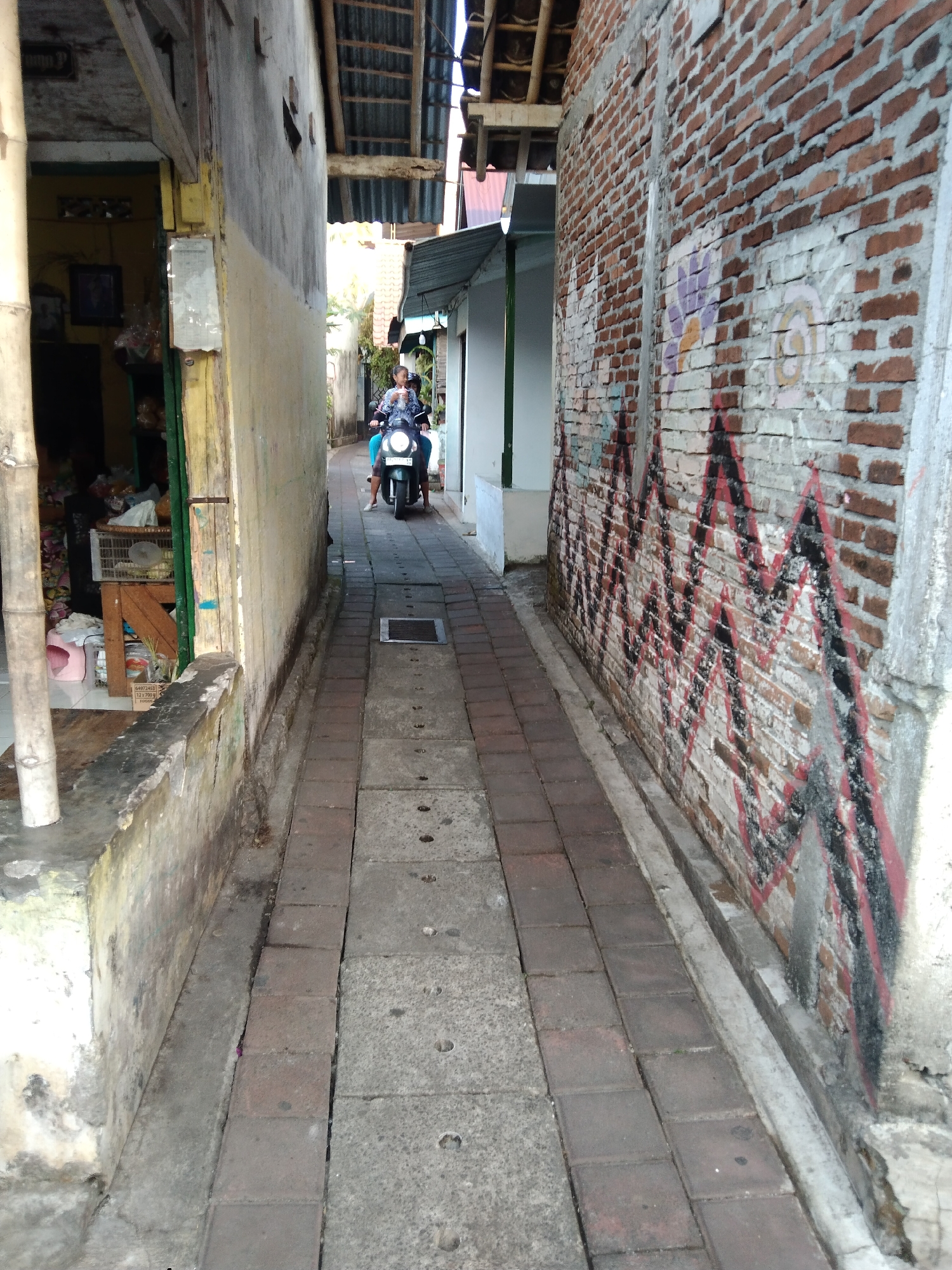 The image depicts a narrow alleyway in the city of Magelang, Indonesia. On the right side, a brick wall is adorned with graffiti featuring a zig-zag pattern in red and black, and some floral designs in purple and yellow. A person is riding a scooter down the alleyway, moving towards the viewer. The scooter is dark green. The alleyway is paved with rectangular bricks. On the left side, there is a building with a faded yellow and gray facade, and a shopfront with various items displayed. A large bamboo pole stands vertically on the left side of the alley. The overall scene suggests a typical urban environment in Indonesia, characterized by its narrow passageways, colorful street art, and everyday activities.
