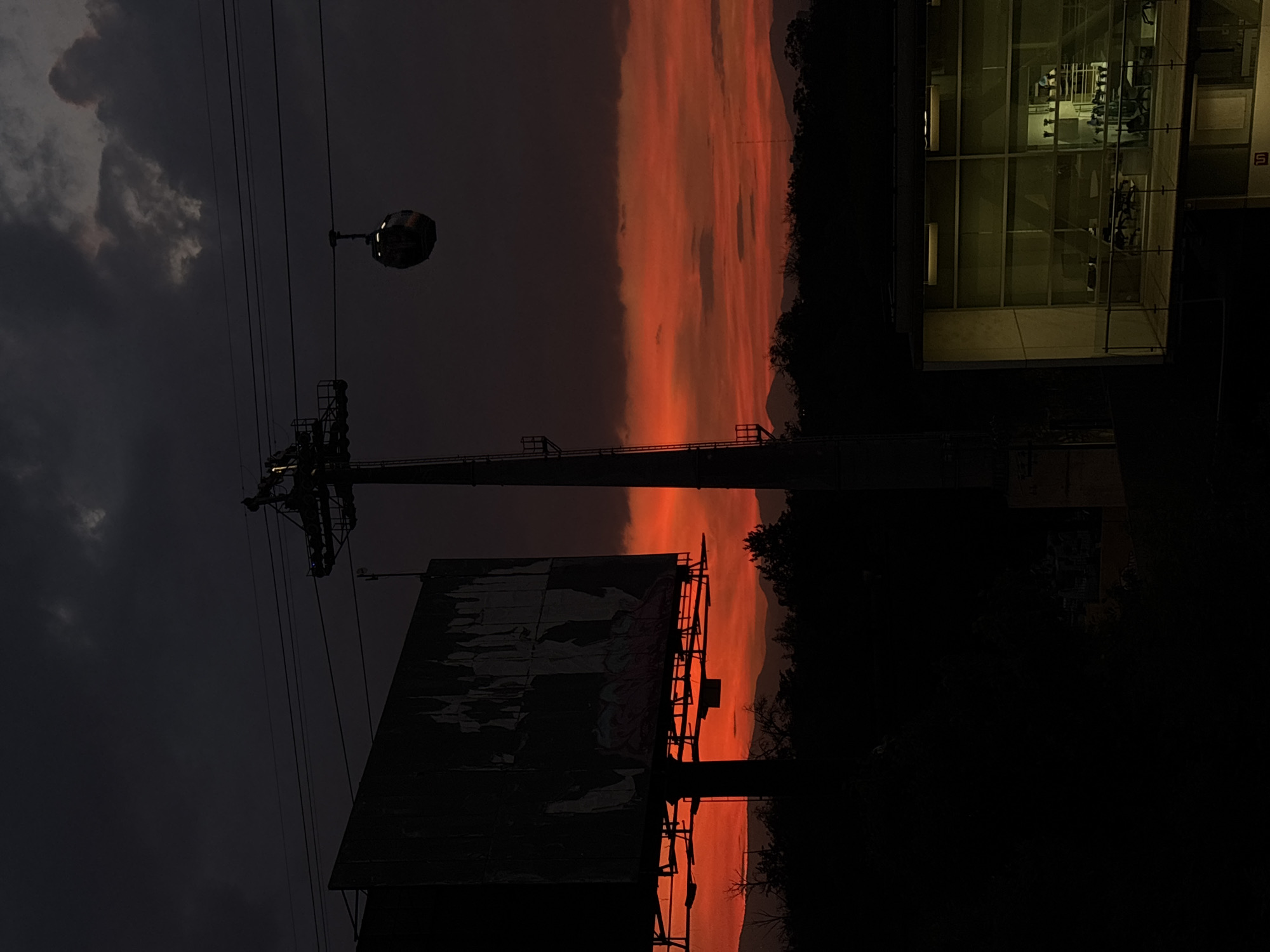 The image depicts an urban scene at dusk or night, likely in Mexico City, Mexico. An intense orange and red sunset dominates the background, obscured by dark trees on the right. In the foreground, a large, weathered billboard stands with visible graffiti. A utility pole with wires and a street lamp are also visible. To the far right, part of a modern glass building is illuminated, showing glimpses of what appears to be a gym inside with exercise equipment. The sky on the left is dark and cloudy, contrasting with the vibrant sunset. The scene is predominantly dark, with light focused on the sunset and the interior of the building.