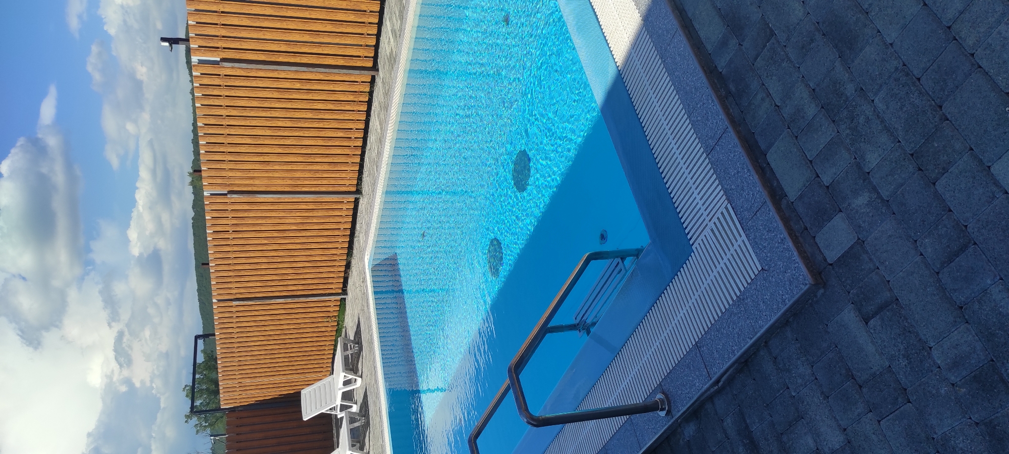 This image displays an outdoor swimming pool with crystal-clear blue water. The pool is surrounded by a paved area made of dark grey interlocking stones, with a wooden slatted fence along one side. The fence is made of horizontal wooden planks and appears to be a light brown color. In the distance, beyond the fence, a lush green landscape with trees is visible, against a bright blue sky filled with white, fluffy clouds.
The water in the pool is inviting and reflects the sky. Several circular features, possibly jets or lights, are visible beneath the surface of the water. A metal handrail leads into the pool, suggesting a step or an entry point. Two white lounge chairs are positioned on the paved area to the left of the pool, indicating a place for relaxation and sunbathing.
The scene suggests a sunny day, likely in the afternoon, given the bright sky and clear visibility. The overall setting appears to be a resort, a private residence, or a recreational area in Tulskiy, Russia. The atmosphere is serene and peaceful, evoking a sense of leisure and enjoyment. There is no visible text in the image.