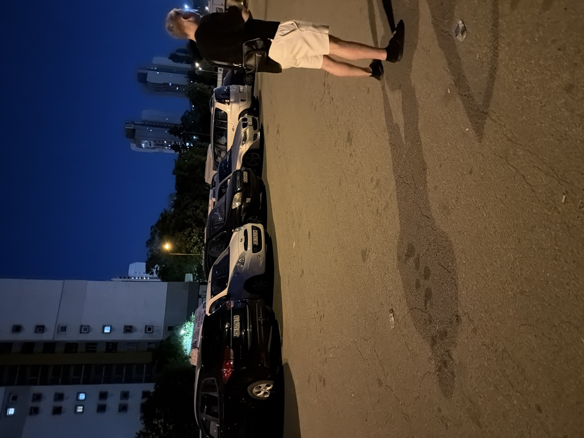 The scene depicts a street in Sofia, Bulgaria at night. A line of parked cars is visible along the right side of the street. A man is standing on the sidewalk, facing the cars. He is wearing a dark shirt and light shorts. The buildings of Sofia, Bulgaria are visible in the background. The sidewalk is littered with cigarette butts and trash.