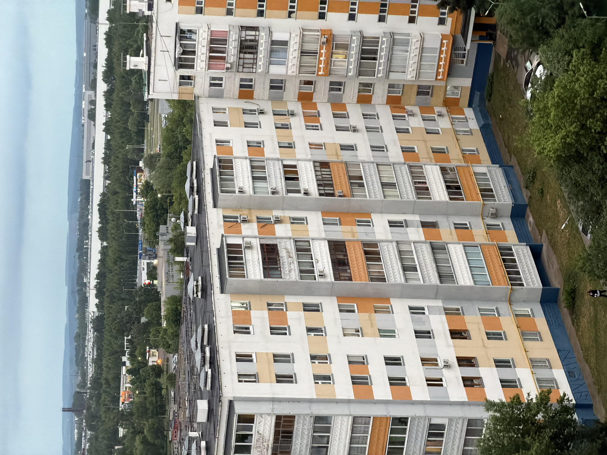 The image shows a view from above looking down on a residential area in Naberezhnye Chelny, Russia. Several tall apartment buildings with a repetitive pattern of white, light brown, and orange facades dominate the view, constructed with balconies and windows. The buildings are surrounded by green vegetation and the edge of a paved area where a car is parked and a person is walking. Beyond the buildings and trees is a road with traffic, and further in the distance, there is a body of water and the horizon. The sky is overcast.