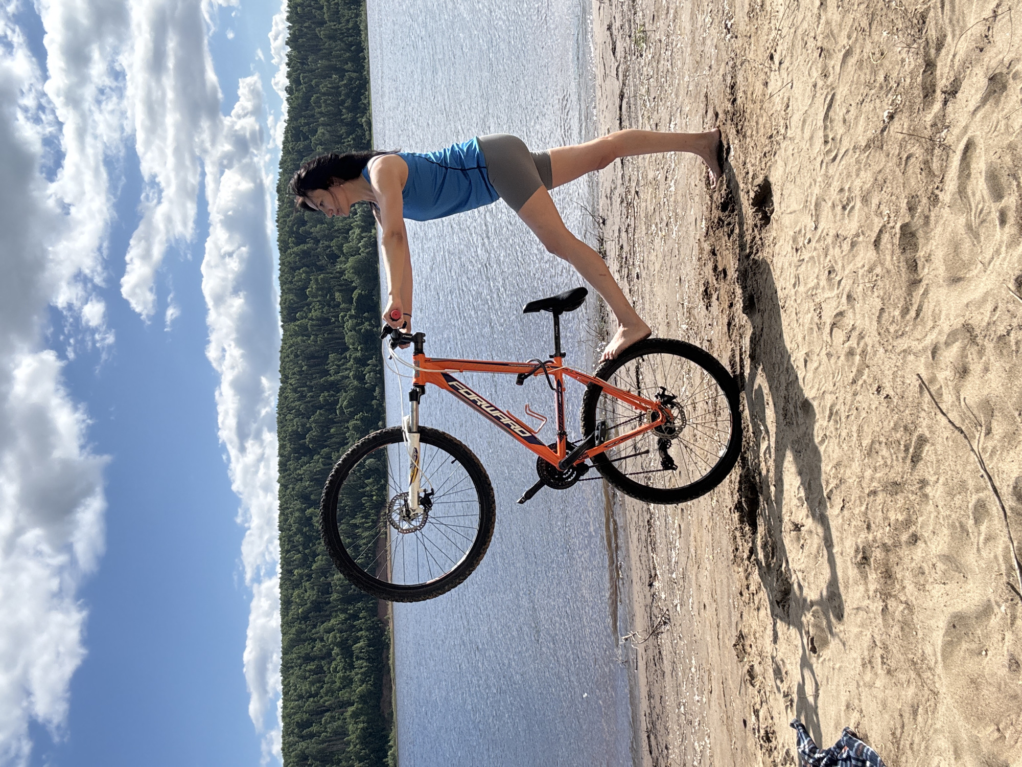 A young woman is performing a yoga pose on a sandy beach next to a lake. She is balancing on her hands with her body extended in a plank-like position, with one leg lifted in the air. An orange and black mountain bike is positioned beneath her, with her hands on the handlebars and her front wheel supporting her upper body weight, while her back wheel is on the ground. The woman has dark hair and is wearing a blue tank top and grey shorts. She appears focused and serene as she holds the pose.
The scene is set on a sunny day, indicated by the bright light and clear blue sky with fluffy white clouds. The lake in the background is calm and reflects the sky. A dense green forest lines the opposite shore of the lake, suggesting a natural, somewhat remote location. The sandy beach has footprints and some dry vegetation scattered across it. The overall atmosphere is peaceful and suggests an outdoor recreational activity.
The visible text on the bicycle frame reads "FORWARD". The image captures a moment of strength, balance, and connection with nature.