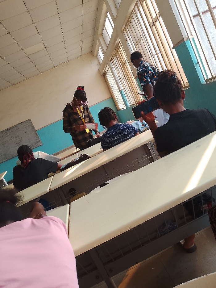 The image shows a classroom scene at an educational institution in Ogboko, Nigeria. A group of students are seated at rows of desks. One student, standing at the front of the classroom, is wearing a plaid shirt and holding a document, possibly addressing the class. Another student is taking a photo using a smartphone. Light streams through a large window with vertical bars. The classroom has light blue walls and a whiteboard.