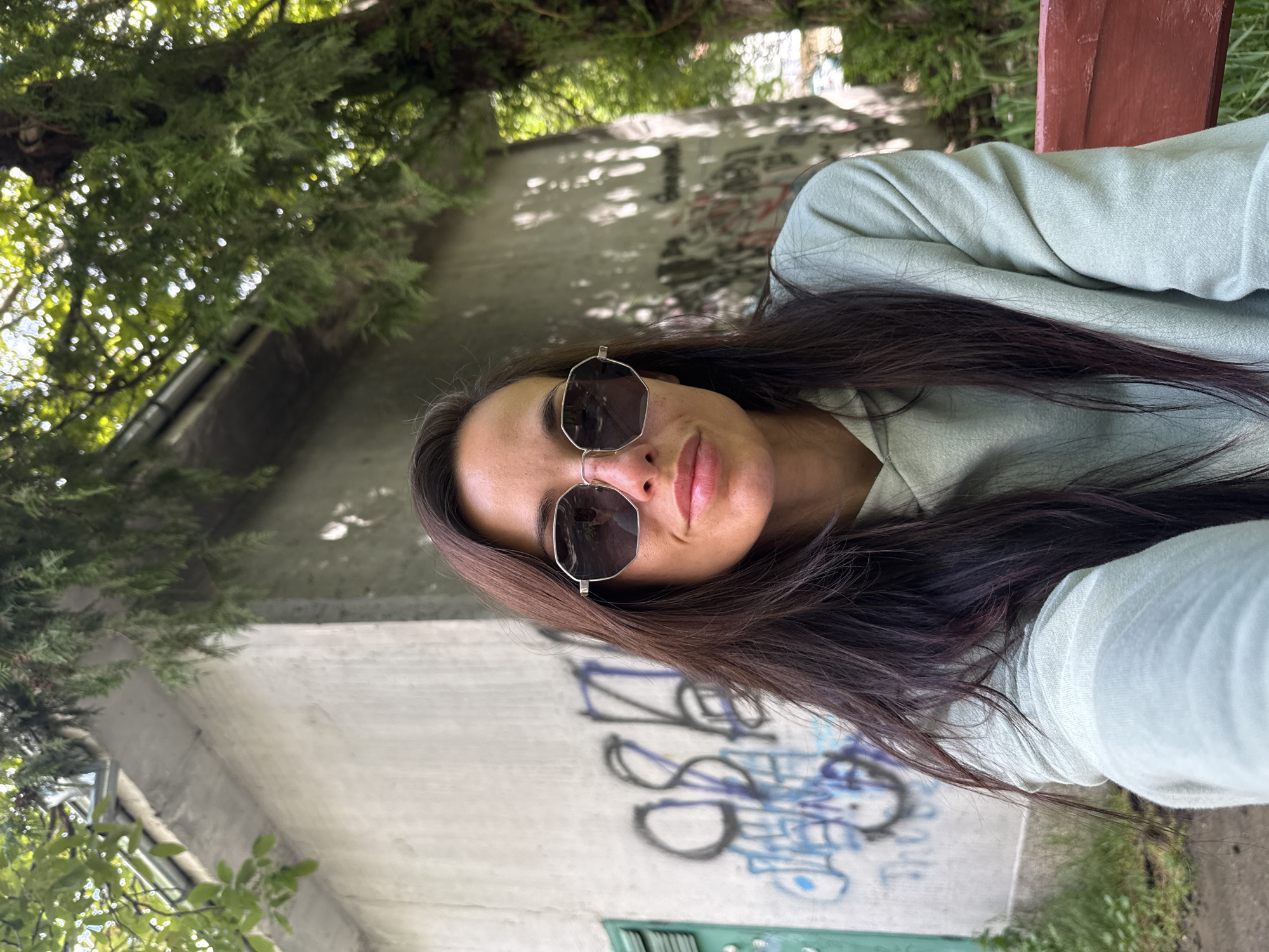 A young adult female is posing for a selfie outdoors, wearing sunglasses and a light green sweater. She is sitting on a bench in front of a concrete structure covered in graffiti. The location appears to be a park-like setting with trees and greenery visible. The specific location in Pleven, Bulgaria, cannot be definitively identified from the image alone.
