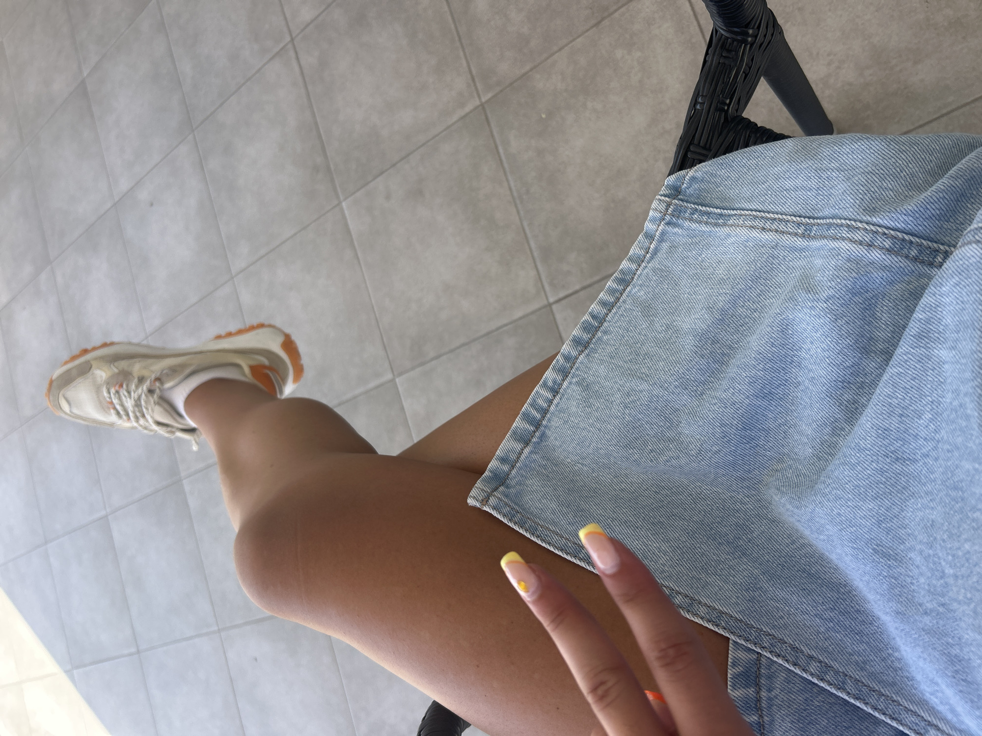 A first-person perspective shot captures a person's legs and lap, adorned with a denim skirt and white sneakers with orange accents. The background reveals a tiled floor, suggesting an outdoor setting, possibly a patio or balcony in Cala Rajada, Spain, given the context. The person's hand is visible in the foreground, making a peace sign with manicured nails featuring yellow tips and a small orange accent. The image is taken from a seated position, with one leg crossed over the other.