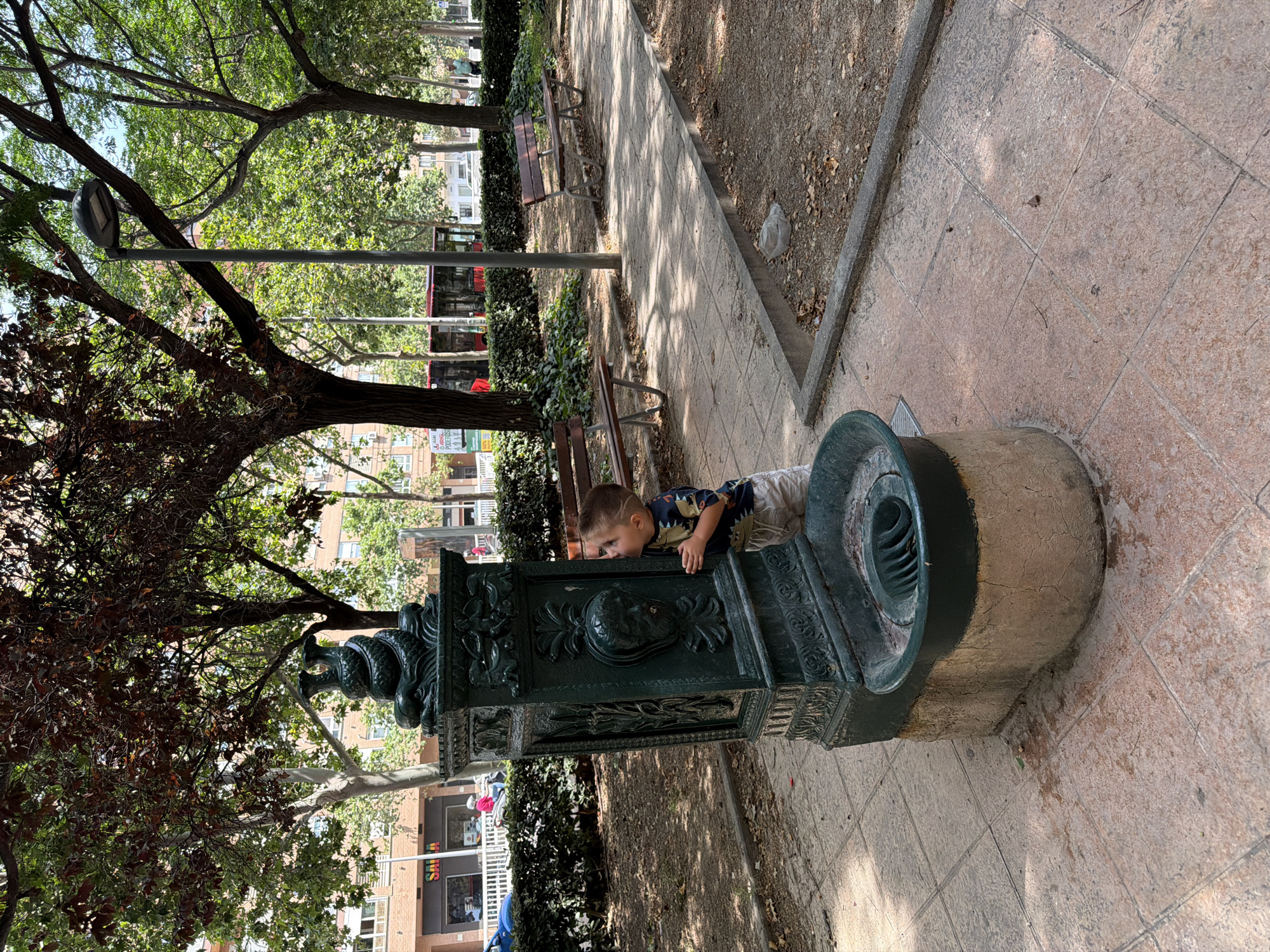 An eye-level shot captures a young child engaging with an ornate public water fountain in a park in Valencia, Spain. The child, with a distinctive shaved hairstyle on the side of his head, wears a patterned dark short-sleeve shirt and light shorts. He is leaning over, looking intently into the basin of the dark green, possibly cast-iron, fountain.
The fountain is elaborately decorated with intricate relief carvings, including faces or masks, foliage motifs, and what appear to be stylized serpents or dragons. Its base is a sturdy, light-colored stone. The setting is a bright, sunny day, indicated by clear shadows cast on the paved pathway. The path is made of large, square, reddish-brown tiles and is bordered by a concrete curb.
Flanking the path are areas of exposed dirt or mulch with scattered dry leaves and low-growing green ivy or shrubs. Mature trees with abundant green foliage provide shade, and wooden park benches are visible along the pathway. In the background, behind the trees, multi-story urban buildings are visible, along with a red bus and other vehicles on a street. A street light pole stands prominently in the mid-ground. On a sign affixed to a building in the background, the word "SALE" is clearly visible, with "PROGRESOS" also discernible below it. The image conveys a casual, everyday moment in a bustling public space.