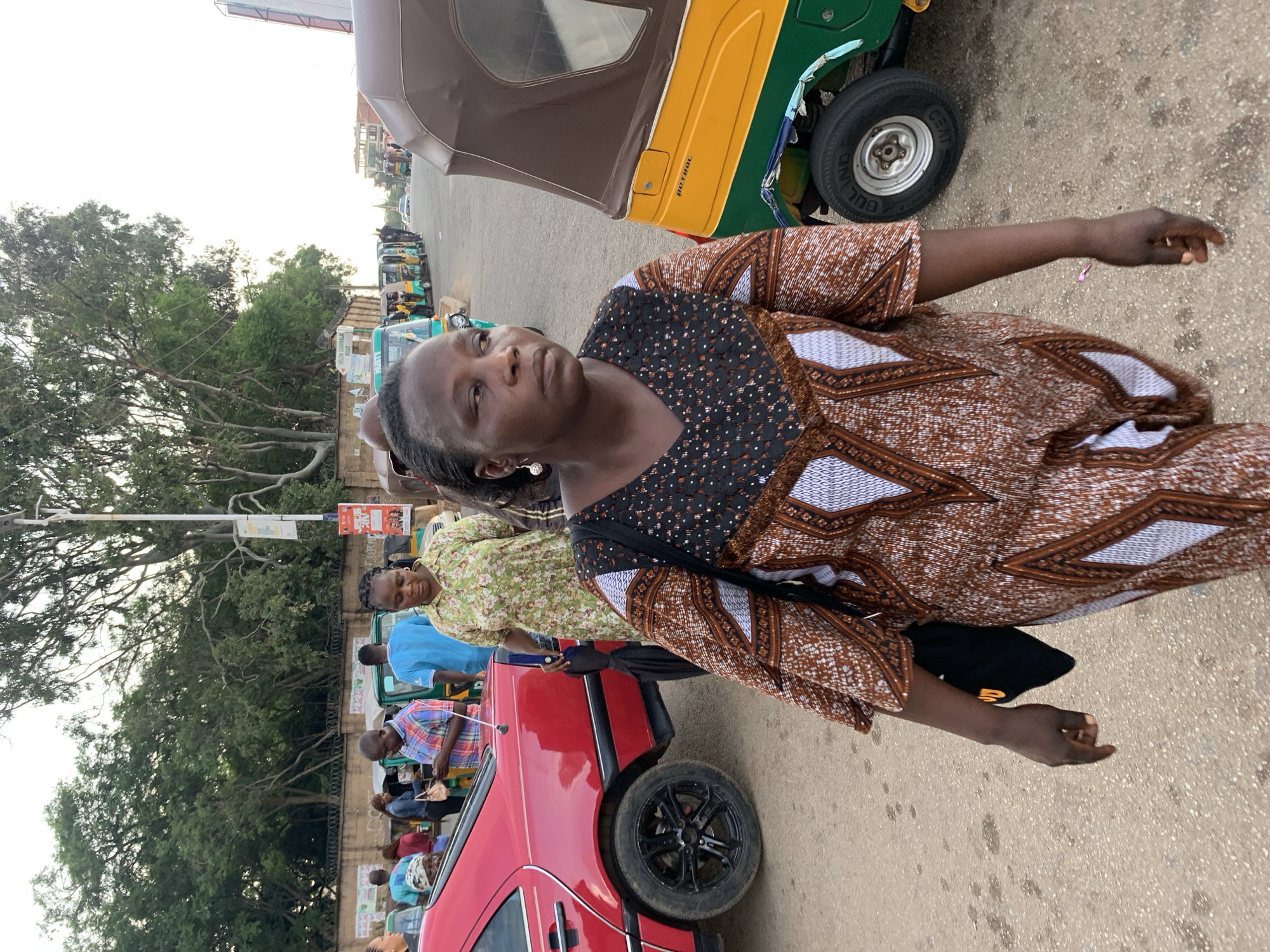 A mid-shot, eye-level, outdoor photograph shows a Black woman walking on a paved road. She is wearing a brown and white patterned dress, and a black shoulder bag. She has a pearl earring in her visible ear. To her right, a yellow and green auto-rickshaw is parked on the road. In the background, other auto-rickshaws, a red car, and several people are visible. Trees and a utility pole are also in the background. The scene appears to be a busy street in Jos, Nigeria.