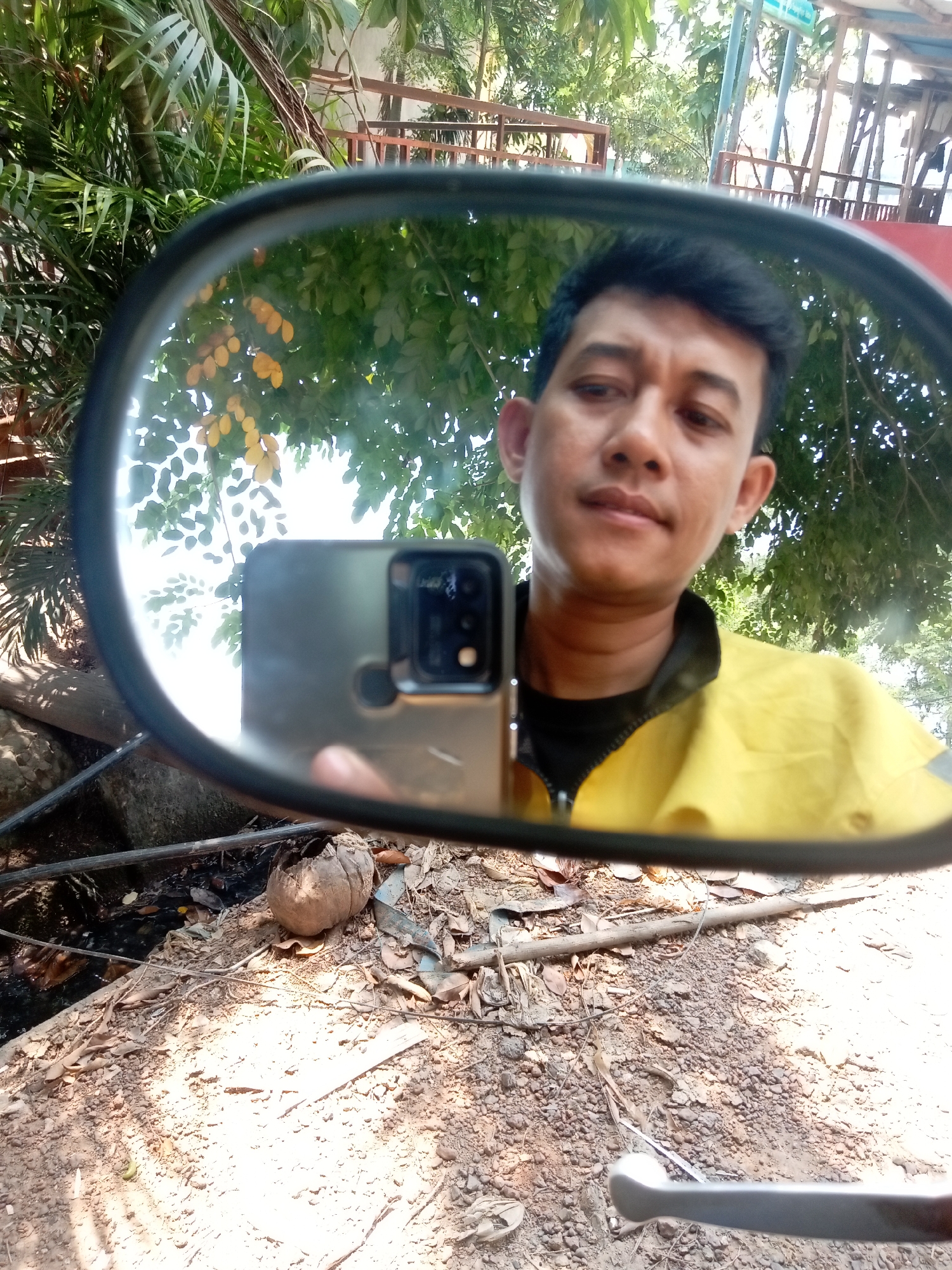 A man is captured in a selfie reflected in a motorcycle's rearview mirror. He is wearing a yellow jacket over a black t-shirt, with short, dark hair. The background visible in the mirror shows lush green foliage, including palm fronds and trees with yellowing leaves. Behind the vegetation, there appears to be a rustic wooden structure, possibly a seating area or building. The foreground outside the mirror's reflection consists of dry, rocky ground with fallen leaves and a discarded coconut shell. The overall scene suggests an outdoor setting, possibly in a rural or semi-rural area within Batam, Indonesia.