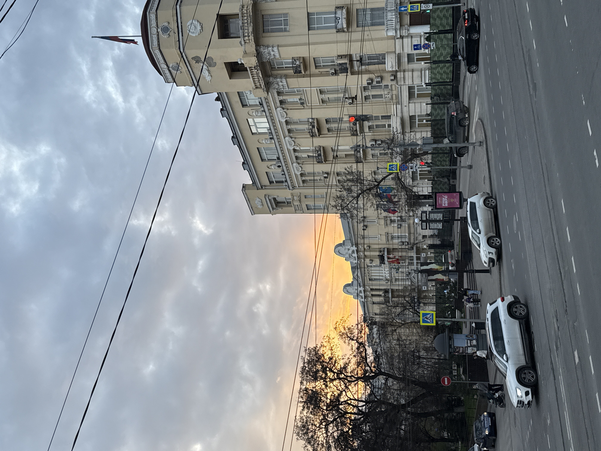 The image shows a street scene in Rostov-on-Don, Russia. The perspective is from an elevated position. The street runs vertically, with a beige building on the left side. This building has multiple floors with windows and ornate architectural details. A Russian flag is on top of the building. Power lines are visible against a cloudy sky, which displays shades of grey and orange. Traffic lanes are marked with white lines. Several parked and moving cars are present, including white and dark-colored vehicles. There are pedestrian crossing signs and crosswalks, indicating areas for pedestrians to cross the street. Bare trees and leafless branches are visible, suggesting it might be autumn or winter. People can be seen walking in the background. A traffic light is visible.