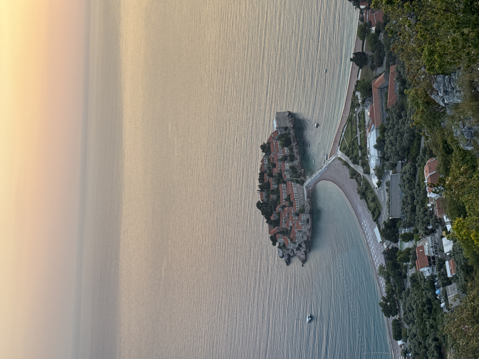The image depicts an aerial view of Sveti Stefan, Montenegro. The island of Sveti Stefan, with its dense cluster of buildings with red-tiled roofs, is centrally located in the frame, connected to the mainland by a narrow isthmus. The Adriatic Sea surrounds the island, showing subtle wave patterns. Adjacent to the isthmus on the mainland is a beach with rows of white sun loungers, indicating a tourist area. Inland from the beach, there is vegetation, including trees and shrubs. Along the coastline, there are buildings, some with red roofs, nestled among the vegetation. The sky appears hazy, particularly towards the left edge of the image, possibly indicating either early morning or late afternoon light.