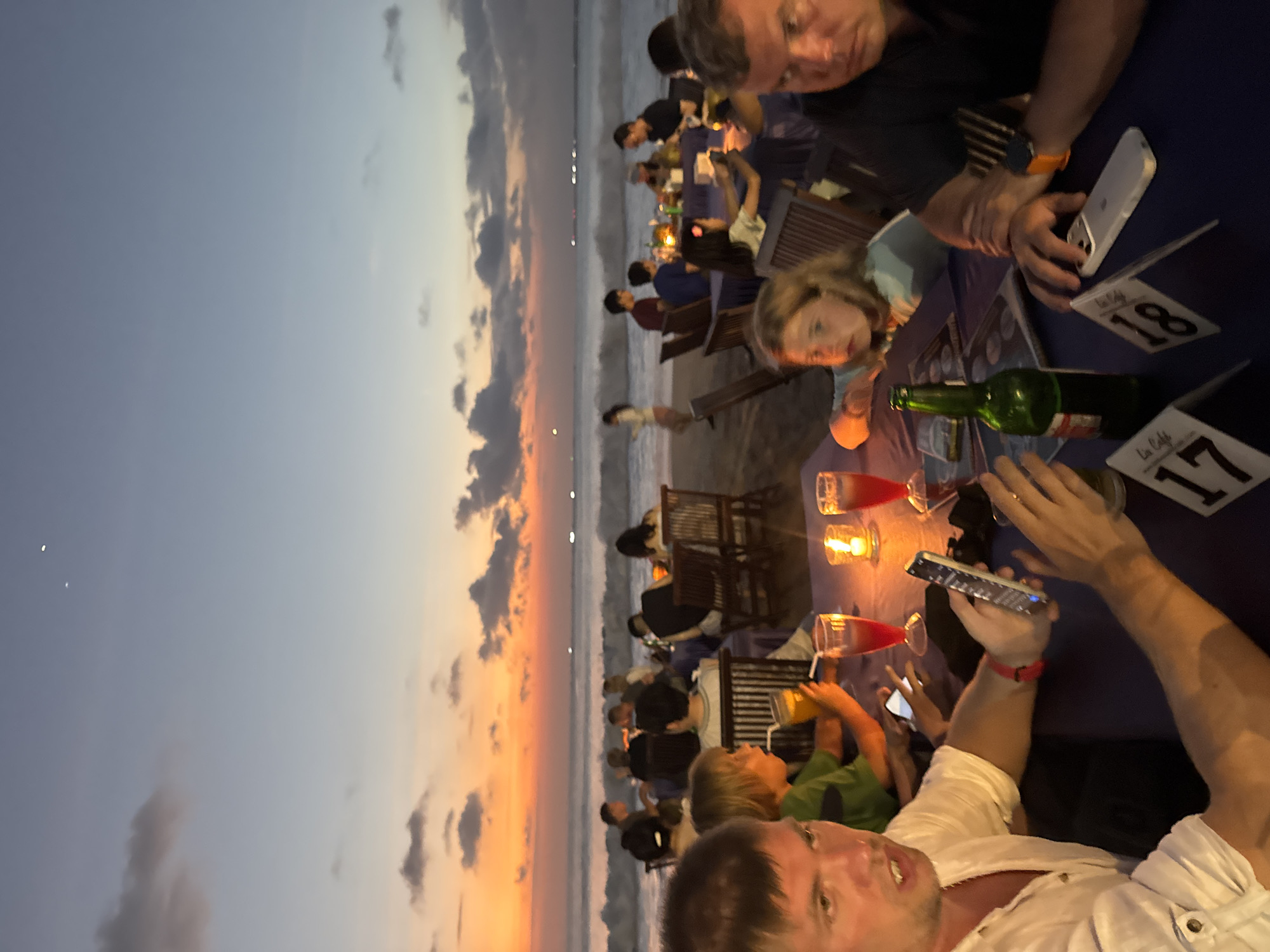 A group of people are gathered at tables on a beach in Kuta Selatan, Indonesia, enjoying a meal as the sun sets over the ocean. The sky is a vibrant mix of orange, pink, and purple, with scattered clouds. The waves are gently rolling onto the shore, and a few distant lights are visible on the horizon. The tables are covered with blue tablecloths and set with glasses, candles, and bottles of drinks. Some people are holding their phones, while others are engaged in conversation. A young girl with blonde hair is looking at the camera, and a man in the foreground is holding up a smartphone. The overall atmosphere is relaxed and convivial.