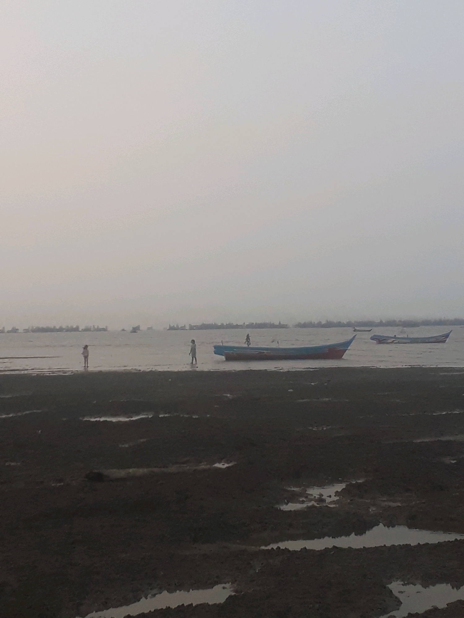 The image depicts a coastal scene in an unspecified Indonesian location, likely during overcast weather. The foreground is dominated by dark, wet mudflats with shallow pools of water reflecting the sky. In the middle ground, a body of calm water is visible, with two small figures, appearing to be children, standing in the shallow water near the shore. Further out in the water, there are several boats. One prominent boat, painted with red and blue accents, is anchored or beached on the mudflats closer to the center of the image. Another boat with a similar color scheme is visible further to the right, partially submerged. In the distance, a blurred line of what appears to be a coastline or a larger settlement is discernible, with numerous boats or structures lined up along it. The overall atmosphere is subdued due to the overcast sky, which creates a diffused light and a muted color palette.