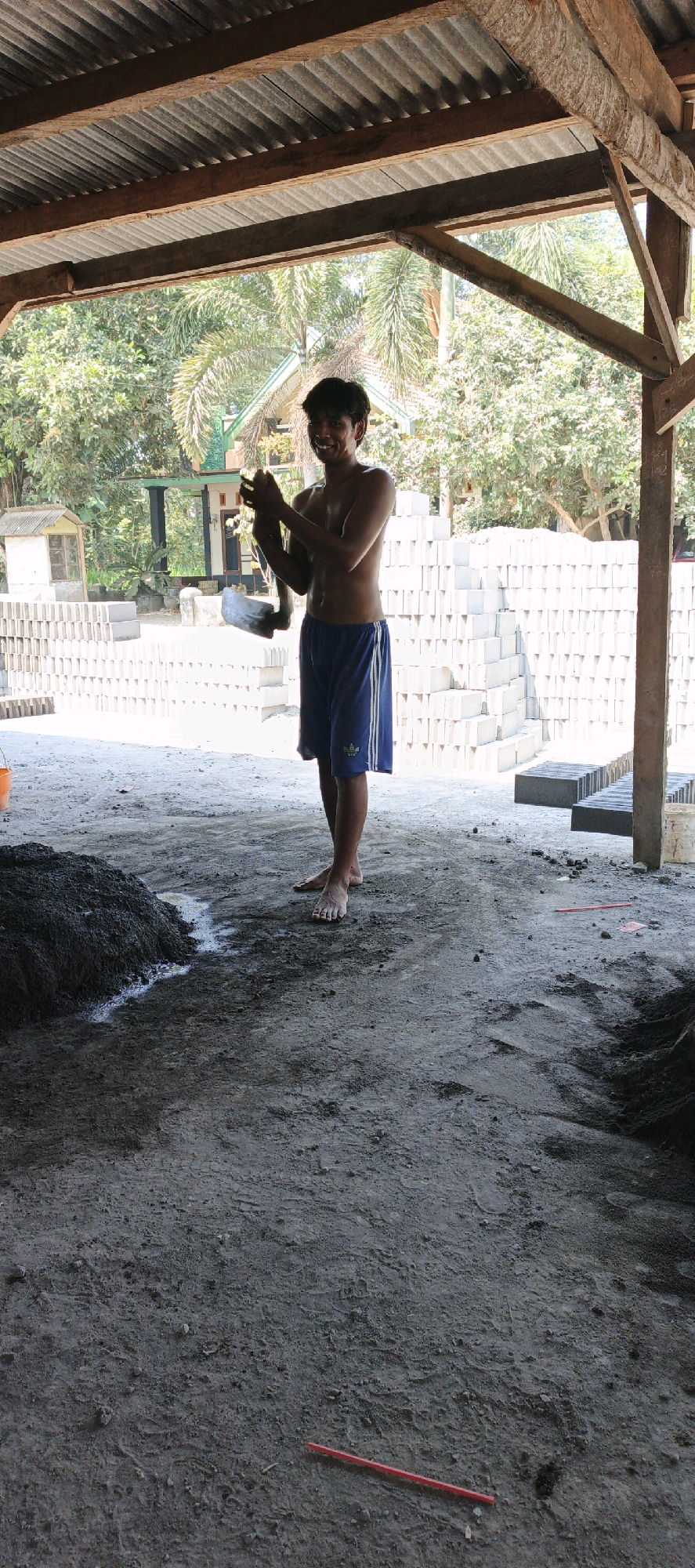 In Kasiyan, Indonesia, a young man stands shirtless under a wooden and corrugated metal roof, smiling and holding a shovel. He wears blue athletic shorts with white stripes. In front of him, a pile of dark, wet material, likely cement or concrete, sits on the dirt ground. Behind him, rows of pale gray bricks are stacked high. The environment appears to be an outdoor worksite, with trees visible in the background. The lighting suggests it is daytime.
