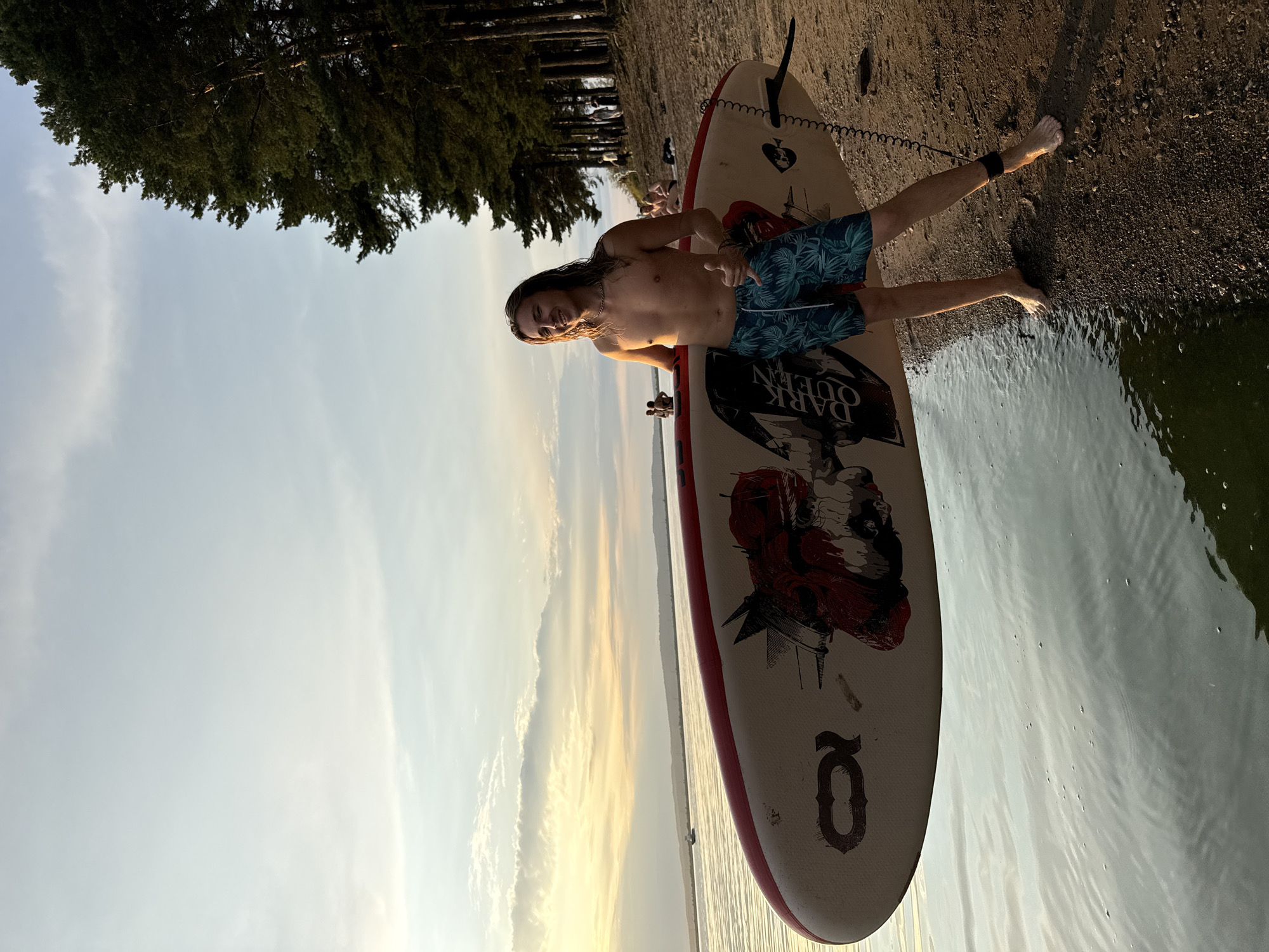 An outdoor medium shot shows a young adult male standing on a sandy beach next to a body of water, holding a paddleboard. He has long blonde hair and is shirtless, wearing blue and green patterned swim trunks. He is smiling and giving a thumbs-up. The paddleboard is white with red trim and a graphic design on it. The water is calm and reflects the sky, which is partly cloudy with the sun setting in the distance. A line of trees is visible on the shore behind him, with a few other people in the background. The lighting suggests it is late afternoon or early evening.