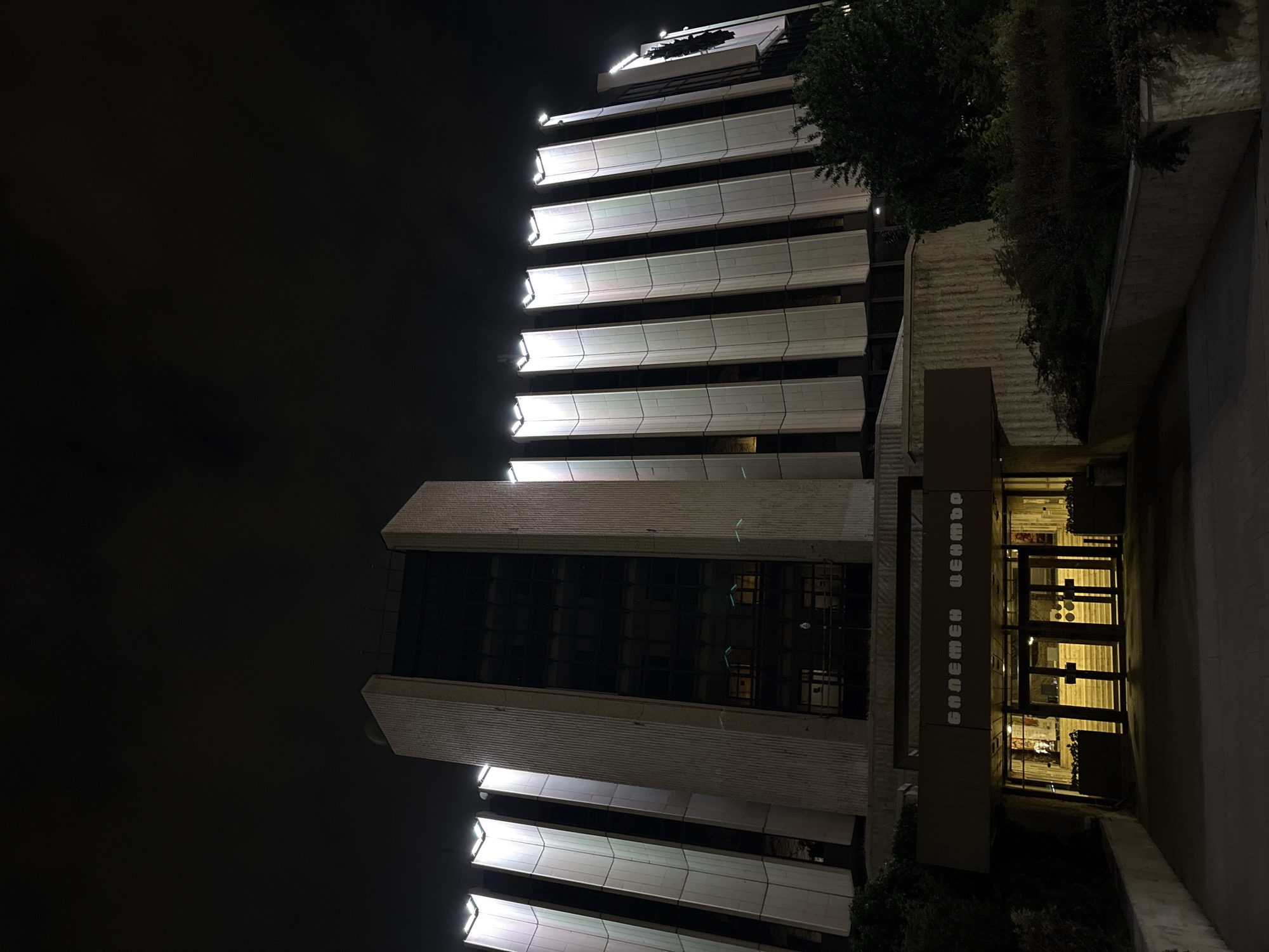 The image shows a tall building at night in Sofia, Bulgaria. The building is lit up with white lights on each floor, which creates a striking visual contrast against the dark sky. The building appears to be a high-rise office building, with many floors and a modern design. In the foreground is the entrance to the building, with a covered walkway and glass doors. The name of the building, "БАНКОВ ЦЕНТЪР" (Bank Center) is vertically displayed. The entrance is brightly lit, illuminating the interior space. The surroundings of the building include some greenery, such as trees and shrubs, near the base of the building. The overall impression of the image is one of a modern urban landscape, with a focus on the architectural features of the building.