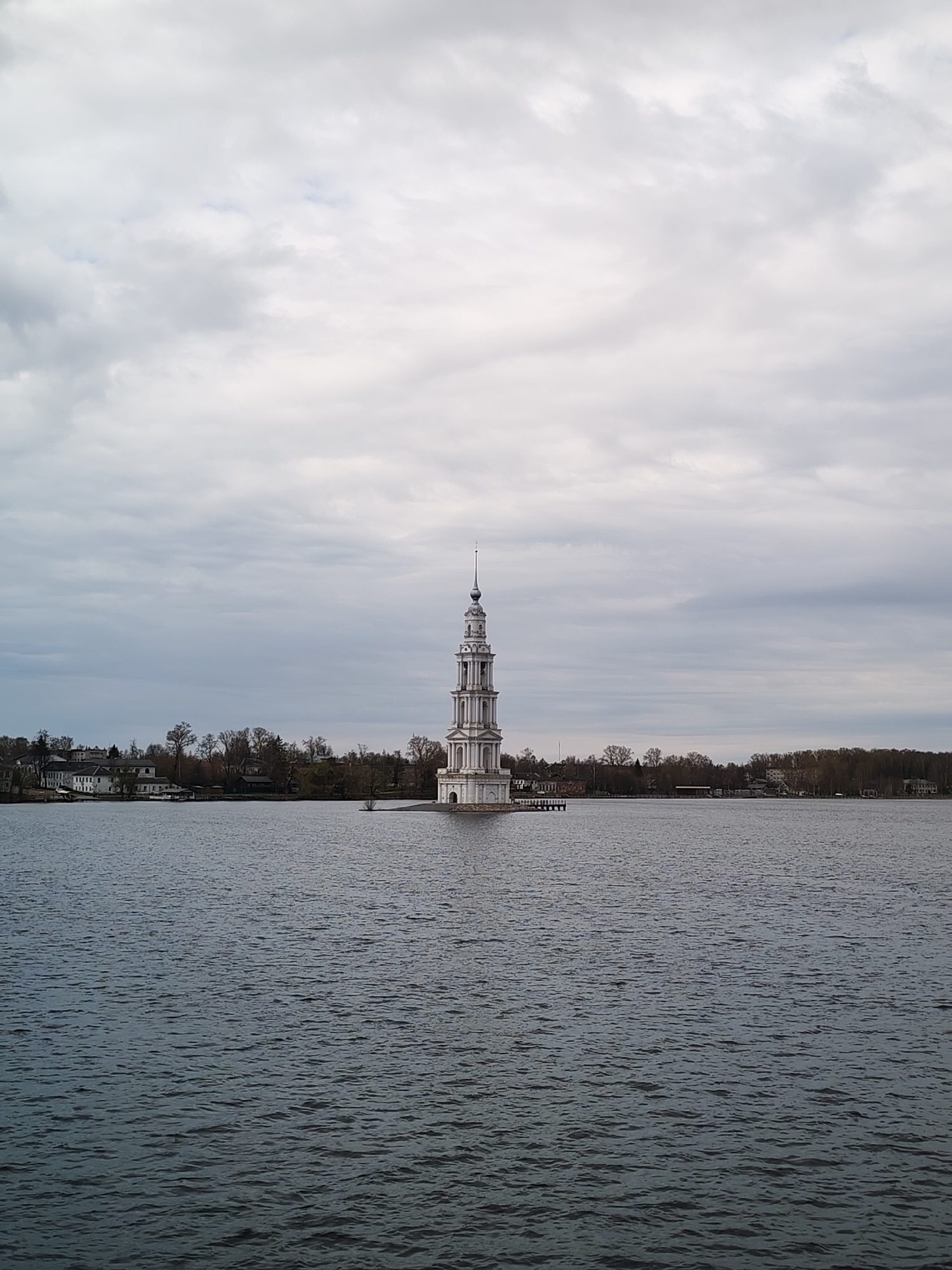 The image shows a free-standing white stone bell tower, the Flooded Belfry of Kalyazin, standing in the middle of a body of water. The tower is the focal point, rising several stories with an ornate design and topped with a spire. The water surface has slight ripples. The skyline behind the tower features low buildings, trees, and a cloudy sky. The scene is located in Kalyazin, Russia, at the site of the Uglich Reservoir. There are no people in the view.