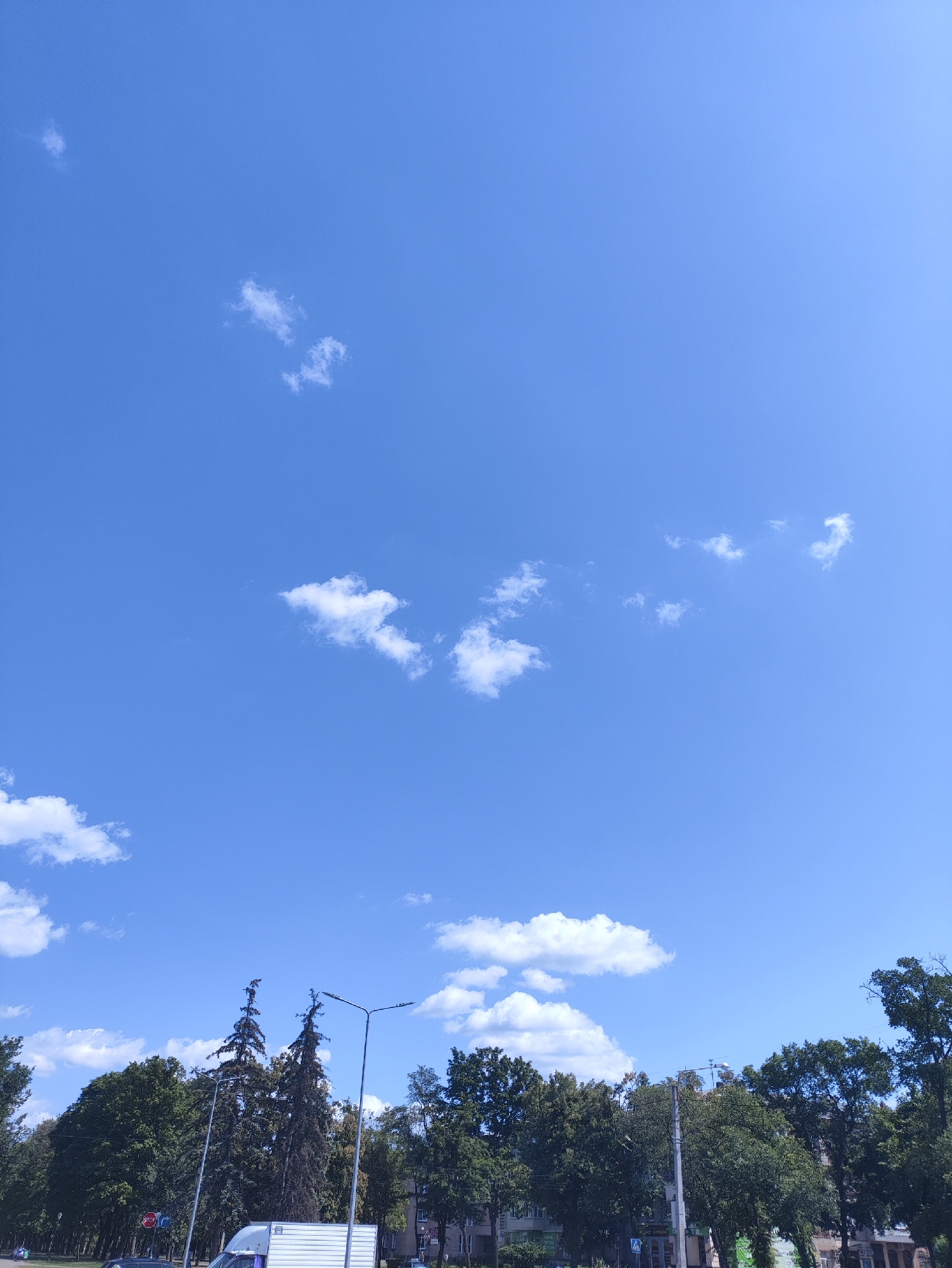 The image depicts a clear blue sky with scattered white cumulus clouds. Below the sky, a line of trees forms a lush green canopy. Several tall streetlights stand amidst the trees, and a white truck is partially visible in the foreground on the left side of the image. A red stop sign is also visible on a pole to the left of the truck. The scene appears to be a street or park in a city, possibly Kharkiv, Ukraine, given the presence of streetlights and urban infrastructure. The overall impression is that of a bright, sunny day.