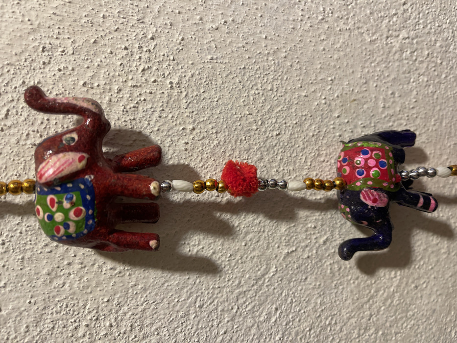 The image captures two ornate, stylized elephant figures connected by a decorative string, positioned against a textured, off-white wall.

The elephant on the left is predominantly reddish-brown, featuring painted white details on its tusks, eyes, and ears. It sports a colorful, saddle-like blanket or covering with distinct blue, green, and white sections, embellished with red and light blue circular patterns. The elephant on the right is dark blue or black, adorned with light pink and white painted features, and a bright red saddle-like blanket decorated with pink, green, and light blue dots. Both elephants appear to be handcrafted, possibly from wood or papier-mâché, showcasing a folk art aesthetic.

A beaded string horizontally links the two elephants. This string is composed of alternating spherical gold-colored beads, spherical silver-colored beads, and elongated white or cream-colored beads. A small, fluffy red pom-pom is centrally located on this string, between the two elephant figures.

The background is a heavily textured wall, likely stucco or rough plaster, in an off-white or cream color. The objects cast soft shadows, indicating indoor lighting. No discernible activity is taking place, as the items are static decorative objects. There is no visible text within the image. While the location context is given as Dosso (Ciappanico), Italy, the image itself does not contain specific visual cues to confirm this.