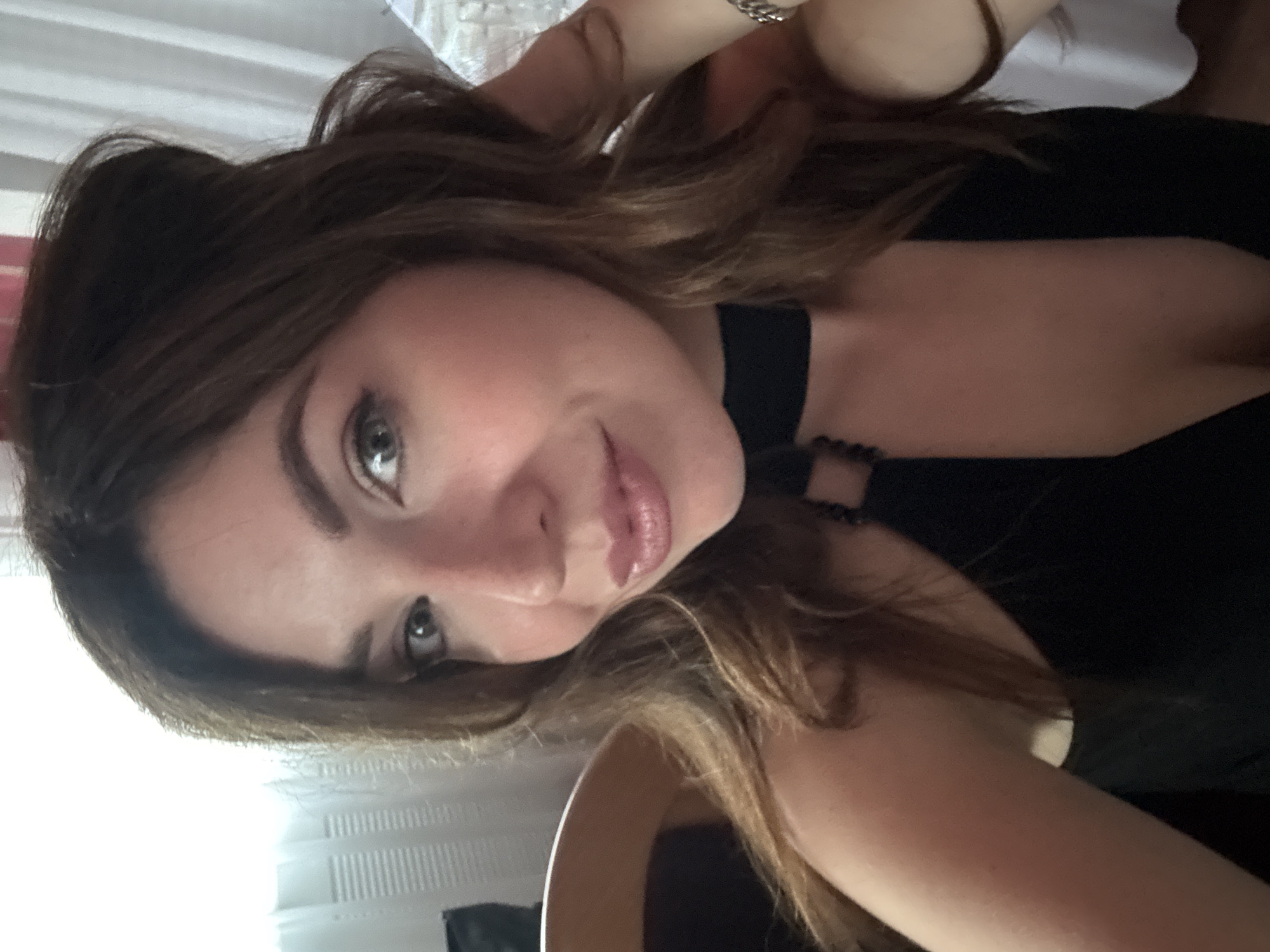A woman with brown hair and green eyes is posing for a selfie in a dimly lit indoor setting. She is wearing a black dress with a choker necklace. The background appears to be a room with curtains and possibly a chair. There is no indication of the city of Kryvyi Rih, Ukraine in the image.