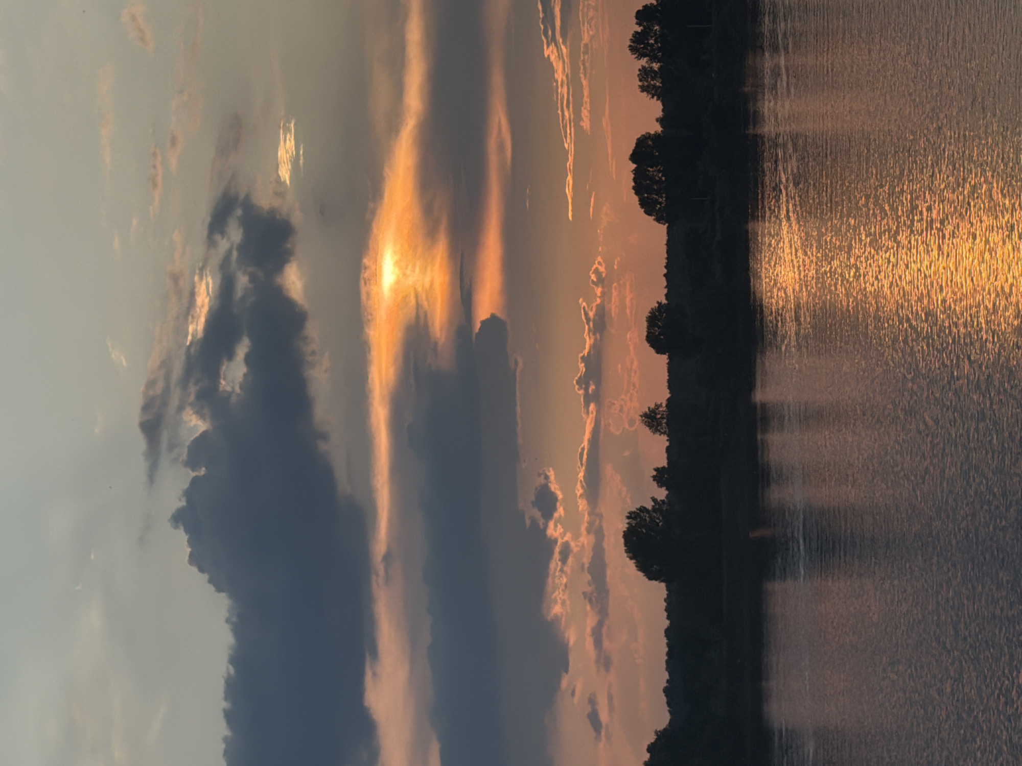 This is a vertical, eye-level, medium-range, outdoor, natural light, full shot of a sunset over the Kama River in Chaikovsky, Russia. The sky is filled with dramatic clouds, some dark and ominous, others tinged with orange and pink from the setting sun. The sun itself is partially obscured by clouds, creating a glowing orange orb. A silhouetted tree line marks the horizon, separating the sky from the dark, reflective surface of the river. The water mirrors the colors of the sky, with streaks of golden light shimmering on its surface. The scene is peaceful and serene, with no people or man-made objects visible.
