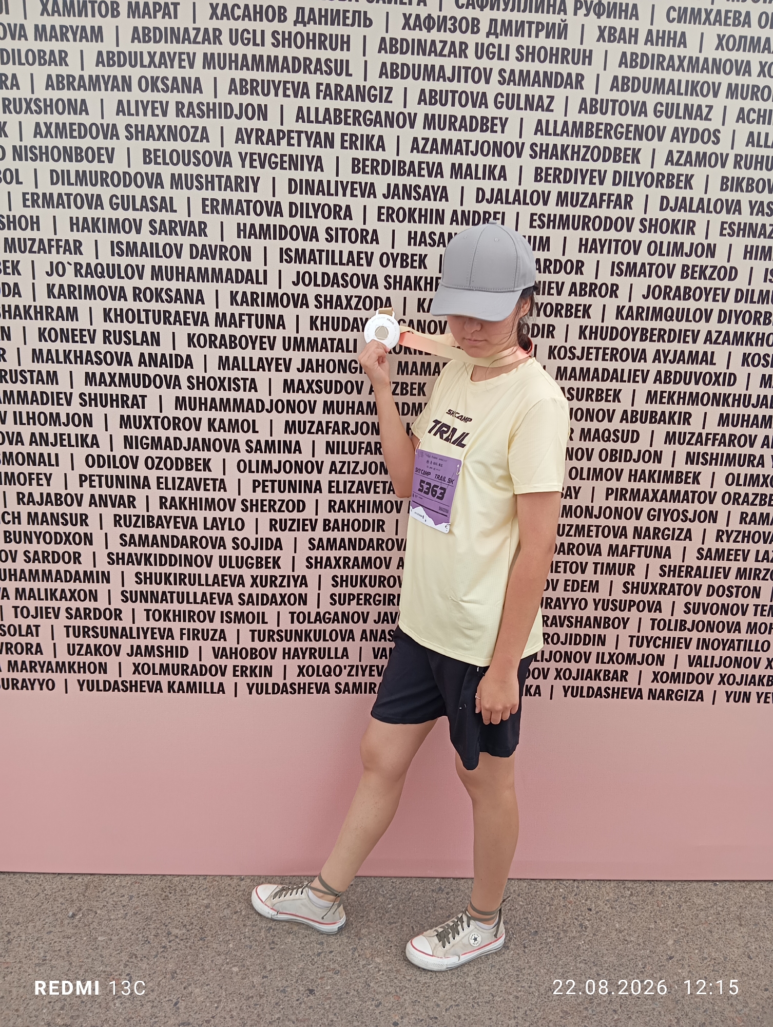 In Amirsay, ski resort, Uzbekistan, a young female, wearing a light yellow t-shirt and dark shorts, stands against a pink backdrop featuring a wall of names. She has a grey cap on her head and is holding a white circular object in her right hand. The names on the wall are printed in black, capital letters, with some Cyrillic and Latin alphabets visible. A white bib with the number 5363 is attached to her t-shirt. The lower part of the image shows a grey, speckled ground and text "REDMI 13C" and "22.08.2026 12:15" in the bottom right corner.