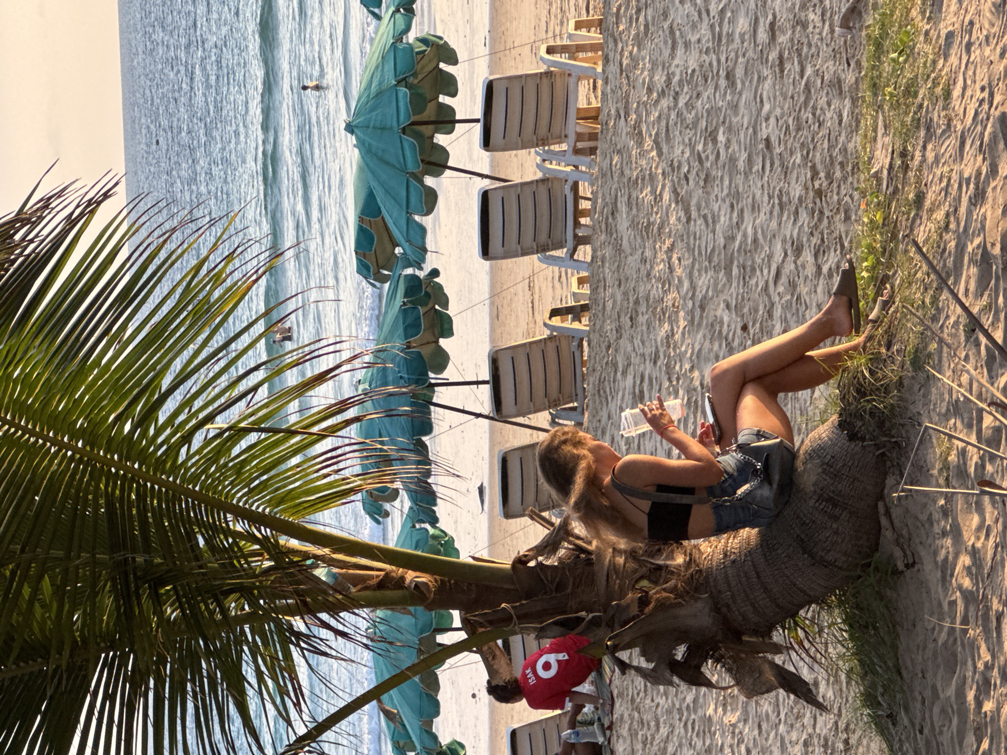 An outdoor, medium shot captures a sunlit beach scene on Karon Beach, Thailand. The foreground is dominated by the base of a palm tree, its trunk at an angle, with a young woman seated on it. She is wearing a black bandeau top and denim shorts, with her left arm resting on her knee and her right hand holding a clear plastic cup. She has blonde hair that falls over her shoulders and is looking down at her cup. Next to her, to the left, is another person, partially obscured, wearing a red t-shirt with the number "9" and the name "ISAK" on the back. In the middle ground, several empty lounge chairs and teal beach umbrellas are arranged on the sand. The background reveals the ocean with gentle waves and a few people swimming or wading in the water, under a warm, hazy sky. The sand is light brown and textured with small ripples.