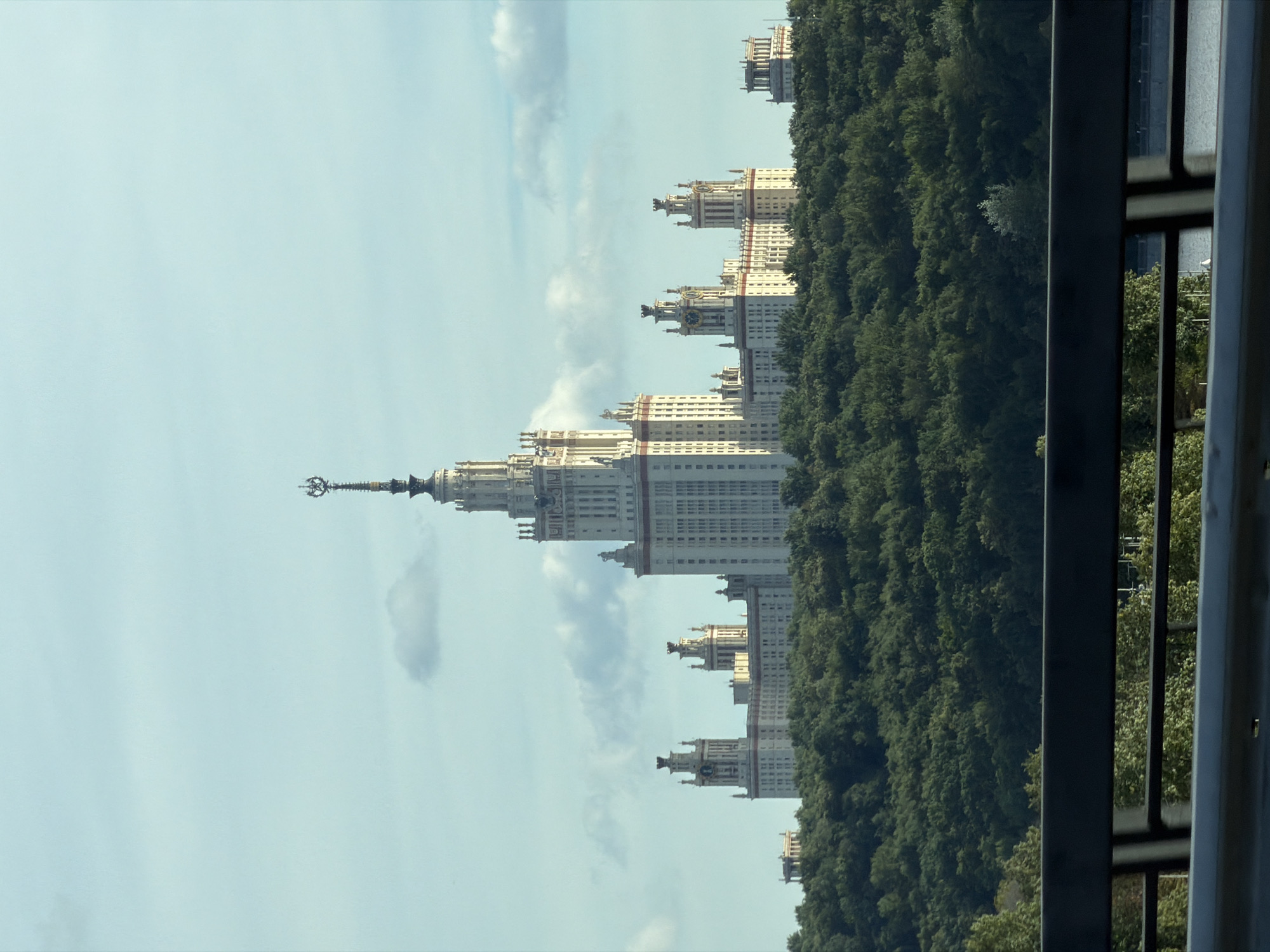 This is a vertical, eye-level, medium-wide shot of the Moscow State University, located in Moscow, Russia. The sky is a pale blue with scattered white clouds. The prominent central building is a tall, cream-colored Stalinist Empire-style skyscraper, topped with a star. To the left of the central tower, there are two smaller, similar-style buildings with prominent clock towers, and further left, a smaller, more classical-style building. The buildings are set atop a densely forested hill. A dark metal railing is in the foreground, obscuring the lower portion of the scene. The overall scene suggests a clear day with good visibility.