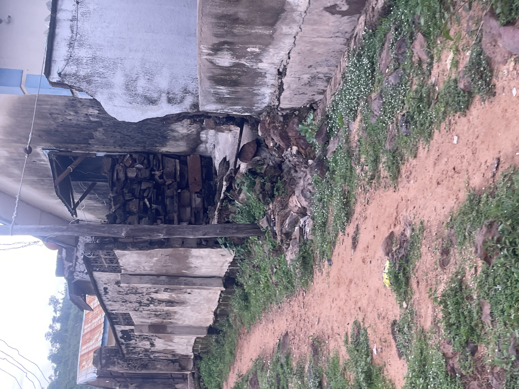 This image depicts a weathered, unpaved pathway running alongside a dilapidated building and a crumbling wall in Lokoja, Nigeria. The pathway is a reddish-brown dirt track with sparse green vegetation and small white wildflowers growing along its edges and in patches on the ground. Large, uneven rocks are scattered on the ground, particularly on the right side of the path.
The building on the right is a multi-story structure with faded, textured walls, some sections appearing to be concrete with visible cracks and water stains. A light blue trim is visible at the top of one section of the building. Adjacent to the building, a pile of stacked stones and bricks are visible, suggesting construction or debris. A wooden post, possibly a utility pole, stands vertically near the wall.
To the left of the pathway is a long, continuous wall constructed of rough, textured concrete. This wall also exhibits signs of age and weathering, with patches of dark discoloration and chipping. It is topped with a metal fence with a decorative pattern. Beyond this wall, a section of a building with a corrugated iron roof, showing signs of rust, is visible. In the background, a lush green hillside covered with trees can be seen under an overcast sky. The overall impression is one of a neglected urban or semi-urban environment in Lokoja, Nigeria.