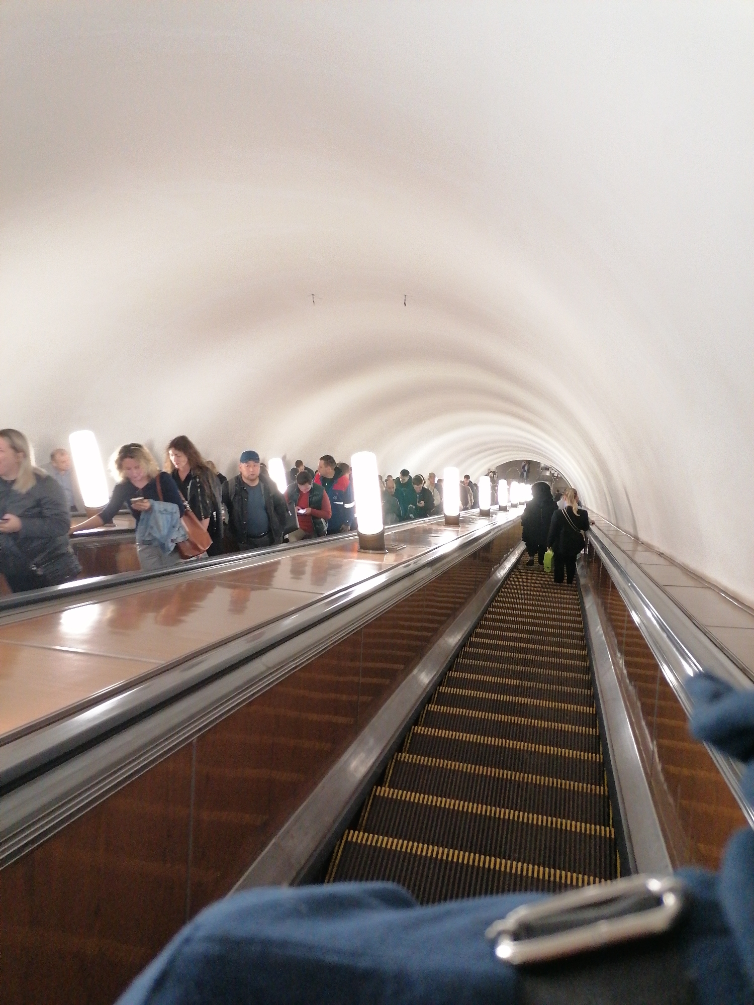The image displays a long, deep escalator tunnel, likely part of an underground subway or metro system. The tunnel has a high, vaulted, off-white ceiling that recedes into the distance, giving a sense of significant depth.
Multiple escalators are visible, running parallel to each other. In the foreground, the perspective is from an escalator going downwards, with its black steps and yellow safety stripes clearly visible. A portion of a blue bag with a metallic clasp belonging to the photographer occupies the bottom center of the frame. The side panels between the escalators are made of a reflective, polished brown material, and gray handrails run alongside.
Numerous people are present on the escalators. On the left side, a line of individuals appears to be traveling upwards, some facing forward, others looking slightly to the side. These commuters are illuminated by several tall, cylindrical light fixtures strategically placed along the escalator route, casting bright white light. Further down the tunnel, more people can be seen, mostly from behind, continuing the stream of passengers. Most individuals are dressed in casual clothing, including jackets and coats, suggesting a typical daily commute.
The scene conveys the atmosphere of a busy urban transportation hub. The artificial lighting inside the tunnel makes the actual time of day indeterminate. No discernible text is visible within the image.