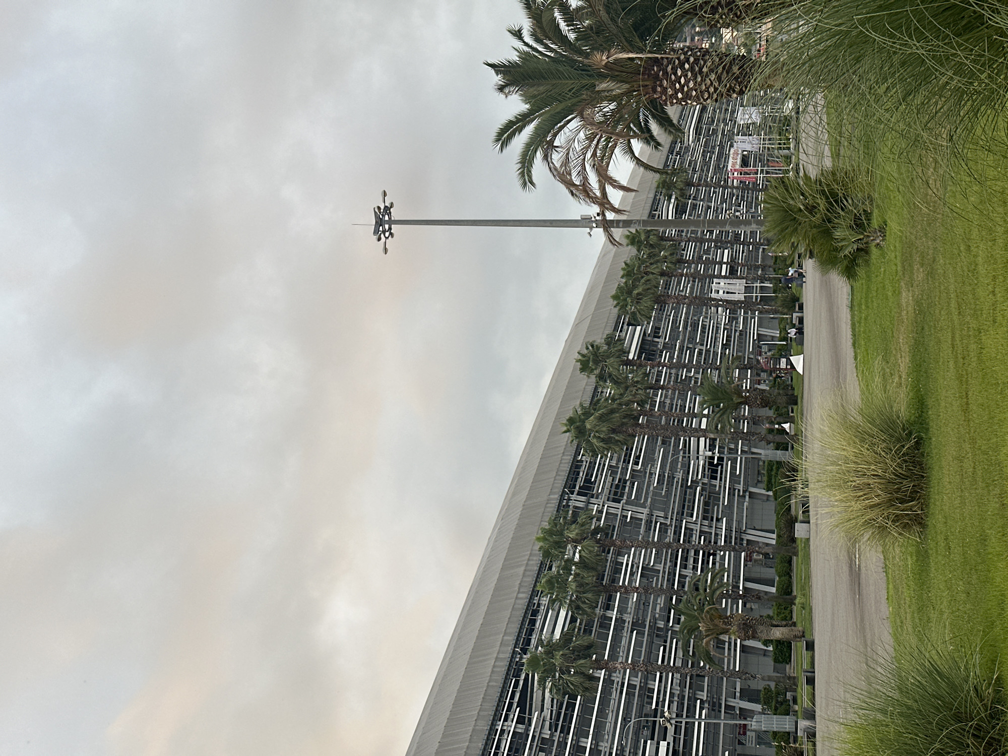 The image shows a modern architectural structure with a white angled roofline and a facade composed of numerous horizontal white elements. Palm trees are planted in front of the structure, with some reaching higher than the facade. A tall pole with multiple lights or antennas stands to the left of the building, with a palm tree to its right. A paved path runs alongside a grassy area to the right of the building, bordered by tall, wispy grass.
In the background, beyond the palm trees and facade, more of the building is visible, appearing to be a grandstands or stadium structure. There are some vehicles parked in the distance, and a red and white sign with text is visible on one of the upper levels. A few people are walking on the path and in the grassy area. The sky is overcast and appears light grey, suggesting a cloudy day. The scene appears to be an outdoor venue, possibly a race track or sports complex, given the grandstand-like architecture and the presence of palm trees, which could indicate a warm climate or a designed landscape feature. The overall mood is neutral, with no strong emotional cues. There is no clear indication of the time of day, though the overcast sky suggests it might be daytime.