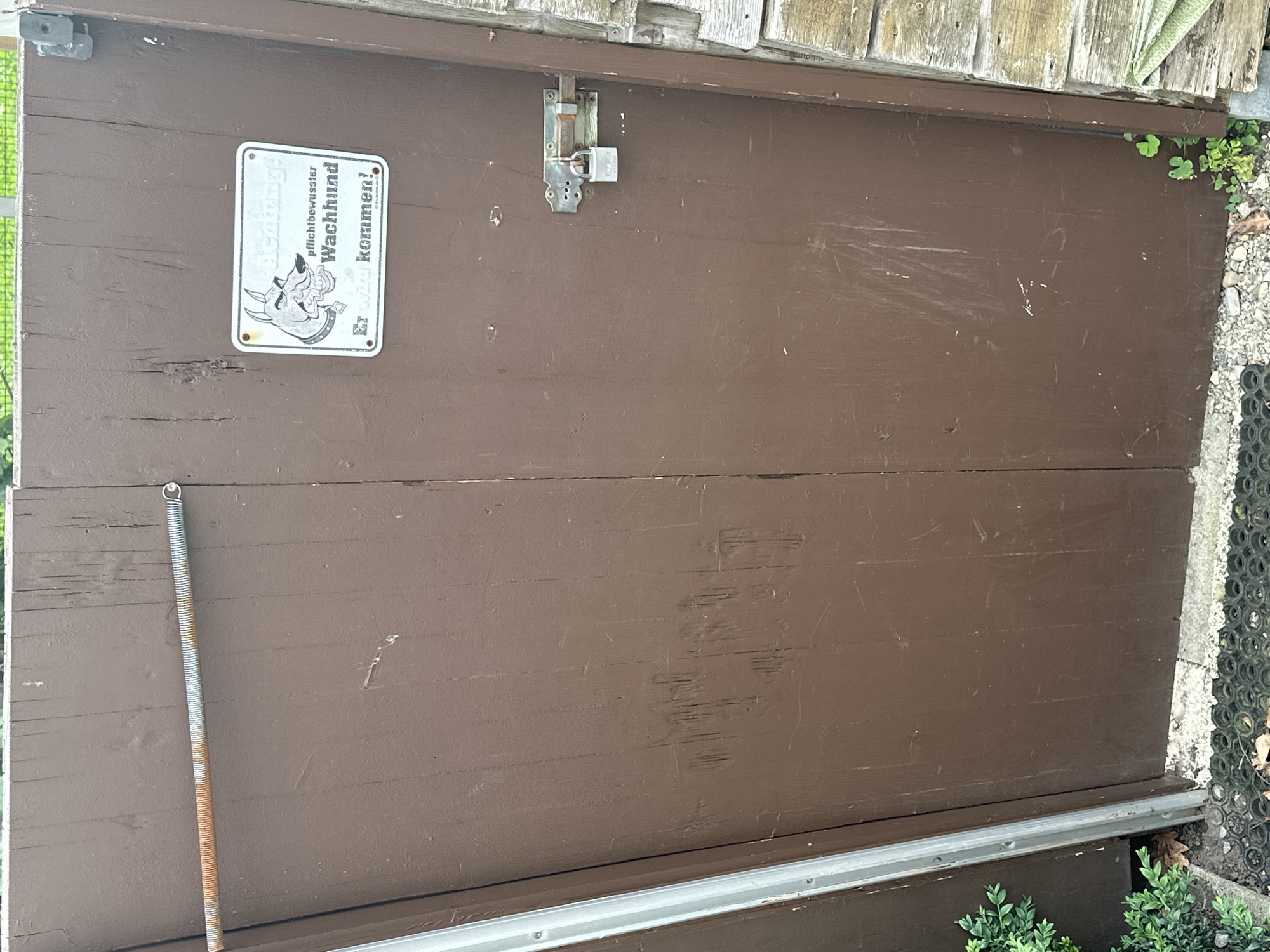 A brown wooden double gate is shown in the image, with a rusty metal spring stretched across the top left portion of the left gate. On the right gate, a white rectangular sign is affixed, depicting a cartoon dog with sharp teeth and the German text "Achtung! pflichtbewusster Wachhund Er wird kommen!". Below the sign, a silver padlock secures a metal latch mechanism to the gate. The gate is surrounded by gravel and some low-lying greenery. The gate appears to be part of a backyard or property boundary in Lauterach, Austria.
