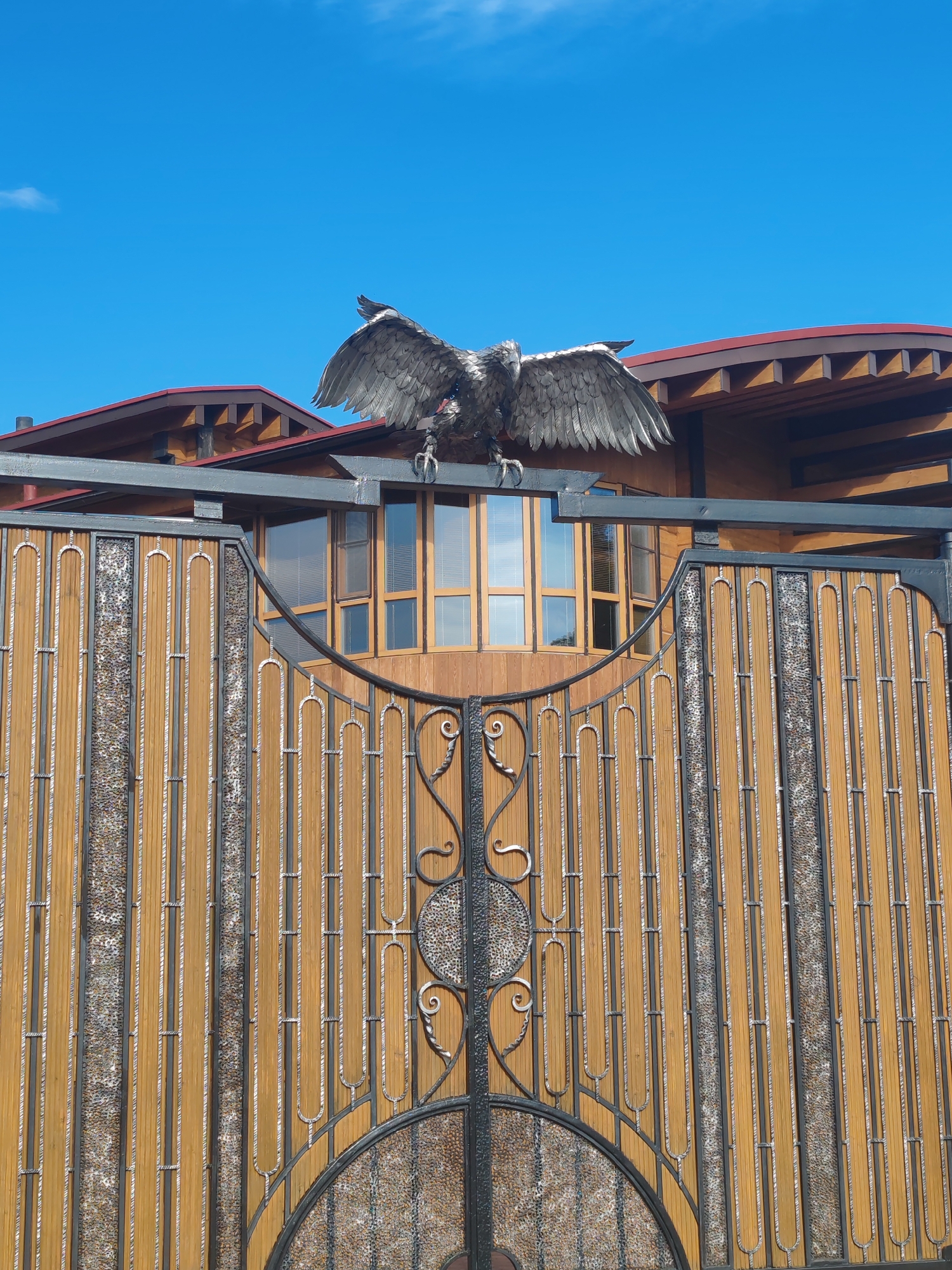 An exterior, eye-level, medium shot captures a detailed view of a decorative metal eagle statue with outstretched wings perched atop a dark metal horizontal bar, which is itself mounted on a wooden and metal gate in Paratunka, Russia. The gate features vertical wooden slats interspersed with textured panels, and intricate black metal scrollwork adorns its lower section. Behind the gate, a building with a dark red roof and a curved wooden facade with multi-paned windows is visible. The sky above is a clear, bright blue with a few wispy white clouds.