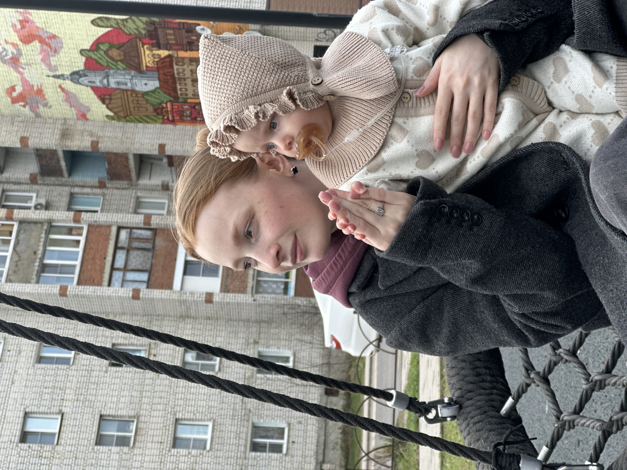 A medium, angled shot captures a woman holding a baby in an outdoor setting. The woman is wearing a dark gray coat over a burgundy-colored hoodie. The baby is wearing a knit hat with a bow and a patterned sweater, and has a pacifier in its mouth. The woman is looking slightly downward with a contemplative expression, and her hands are clasped together. Behind them is a tall building with a mural. The ropes of a playground swing are in the foreground. Based on the presence of the mural, the location is in the city of Pokrov, Russia.