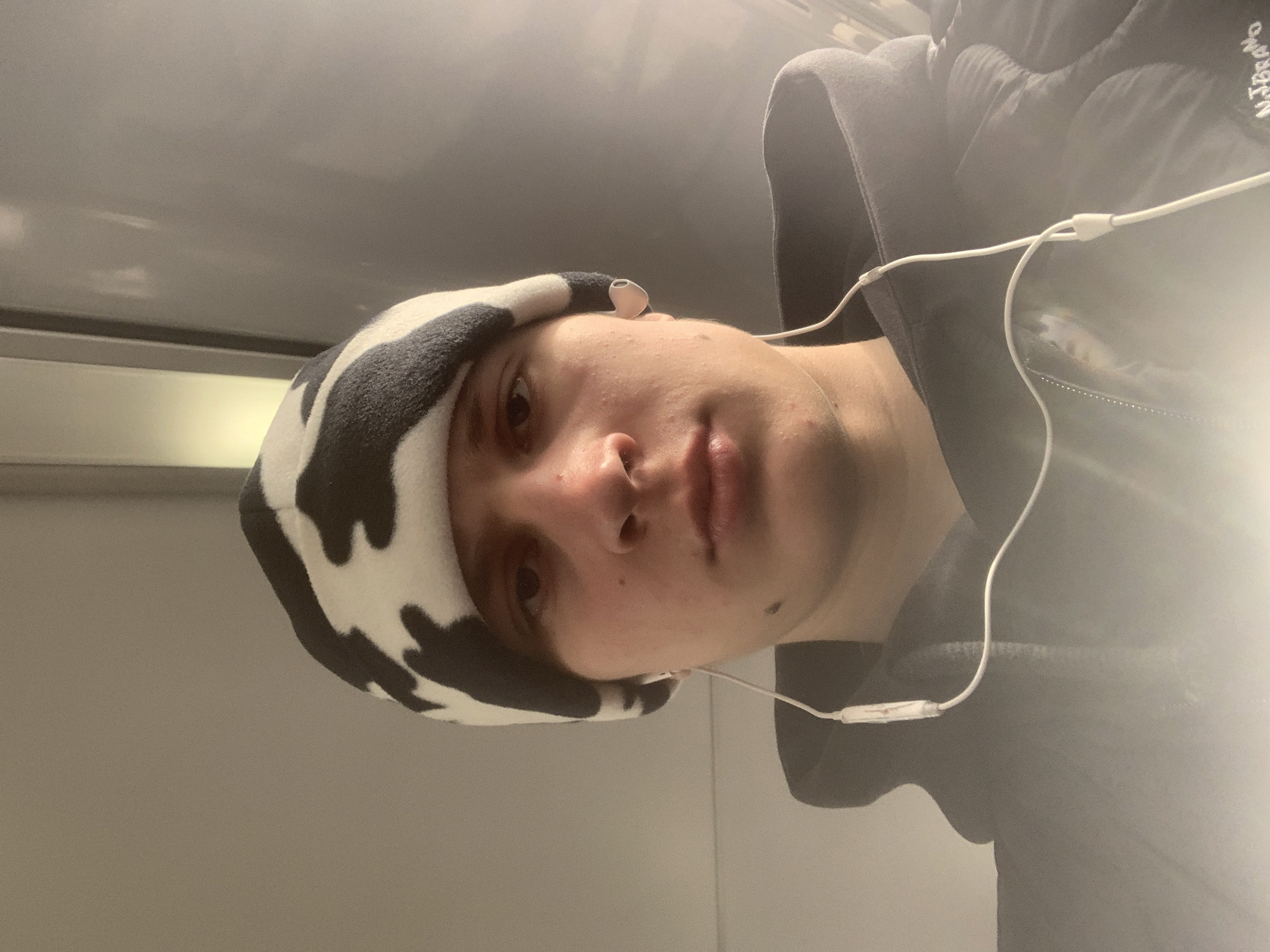 A young individual is depicted in a close-up, head-and-shoulders portrait taken indoors in Krasnoyarsk, Russia. The subject, appearing to be male, is wearing a black and white cow-print beanie hat and a dark-colored hooded garment. White wired earbuds are inserted into both ears, with the wire and an inline control module visible extending down the front of the jacket. The subject has light skin, a prominent nose, and a dark mole or mark on the right cheek (viewer's left). The eyes are open, and the gaze is directed towards the camera. The background consists of indistinct light-toned, possibly paneled, surfaces, consistent with an interior environment, possibly within a vehicle or building interior. Lighting appears to be frontal, illuminating the subject against a somewhat dimmer background. A faint white embroidered text, "N.J BRAÑO", is discernible on the lower right chest area of the subject's jacket.
