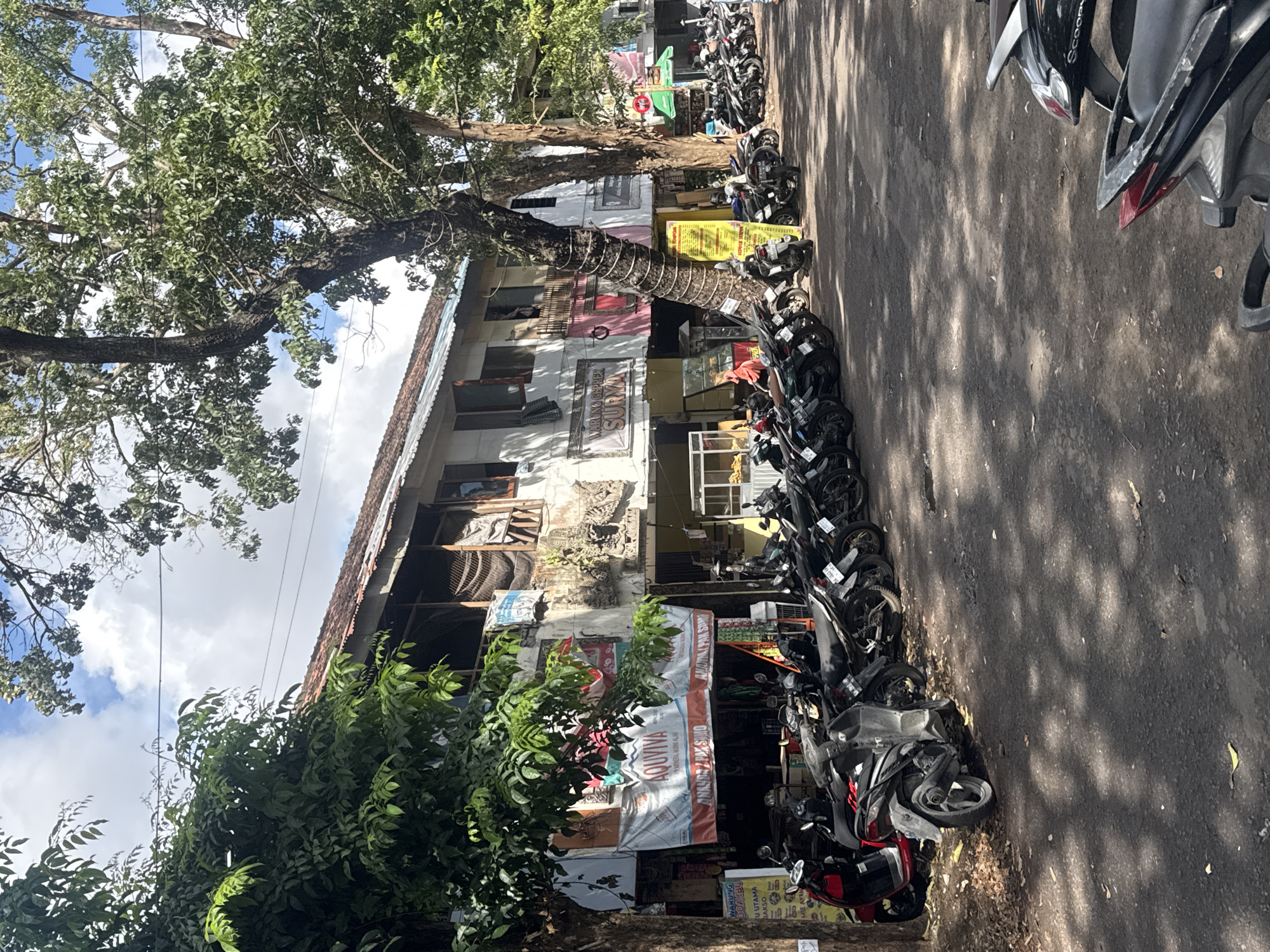 In Denpasar, Indonesia, a row of parked motorcycles lines a paved street in front of a weathered, two-story building. The building's facade is decorated with various banners, one prominently featuring the text "AQUIVA" and another with "WARUNG GEPREK SURYA". Several trees with dense foliage cast dappled shadows across the scene. The sky above is partly cloudy. The area appears to be a commercial street with shops or eateries.