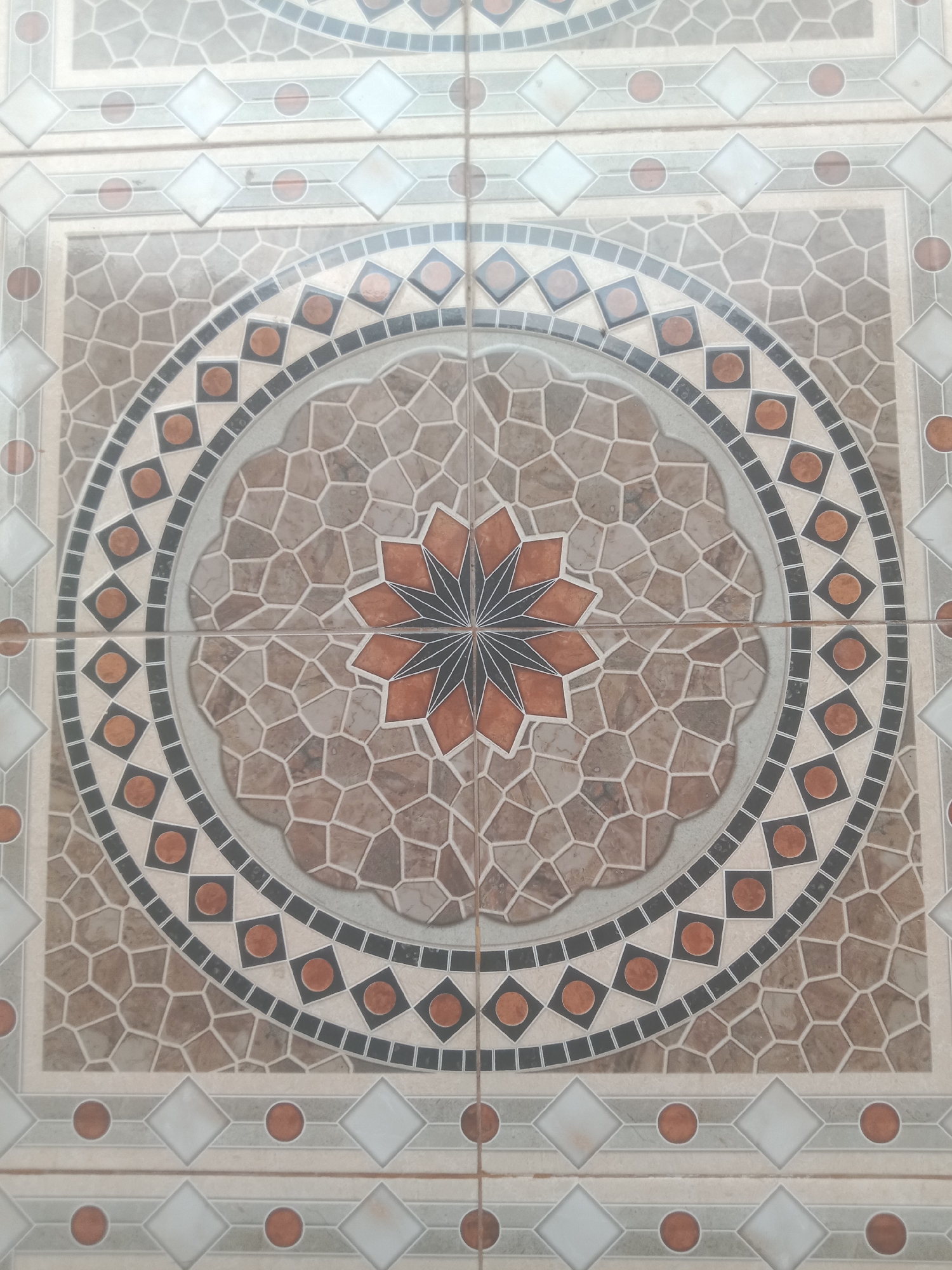 The image shows a close-up, top-down view of a decorative tiled floor. The tiles feature a circular mandala-like pattern in shades of brown, gray, white, and orange. The central element is an eight-pointed star with orange and black accents, surrounded by a textured brown and gray pattern resembling stones. This is enclosed by concentric rings of smaller black and orange squares, and a wider gray band with an undulating edge. The outer border of the circular design is a ring of orange circles within black squares. The surrounding tiles have a simpler geometric pattern of white diamonds and orange circles interspersed with gray and beige lines. The image does not depict any people or identifiable landmarks, and therefore, its location in Kedungsari, Indonesia, cannot be determined from the visual information alone. The scene is an interior or exterior floor surface.