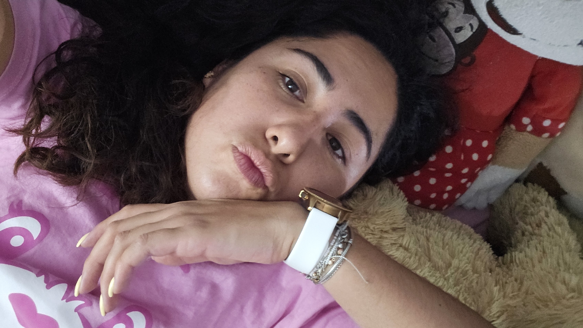 The image shows a woman lying down, possibly in San Bernardino Tlaxcalancingo, Mexico, although the location cannot be definitively determined from the image alone. She is resting her head on her hand, wearing a pink shirt and a gold watch with a white band. Her hair is dark and curly. Several bracelets are visible on her wrist. Next to her are a brown teddy bear and a red and white polka dot stuffed animal, possibly Minnie Mouse. The image has a selfie-style perspective.