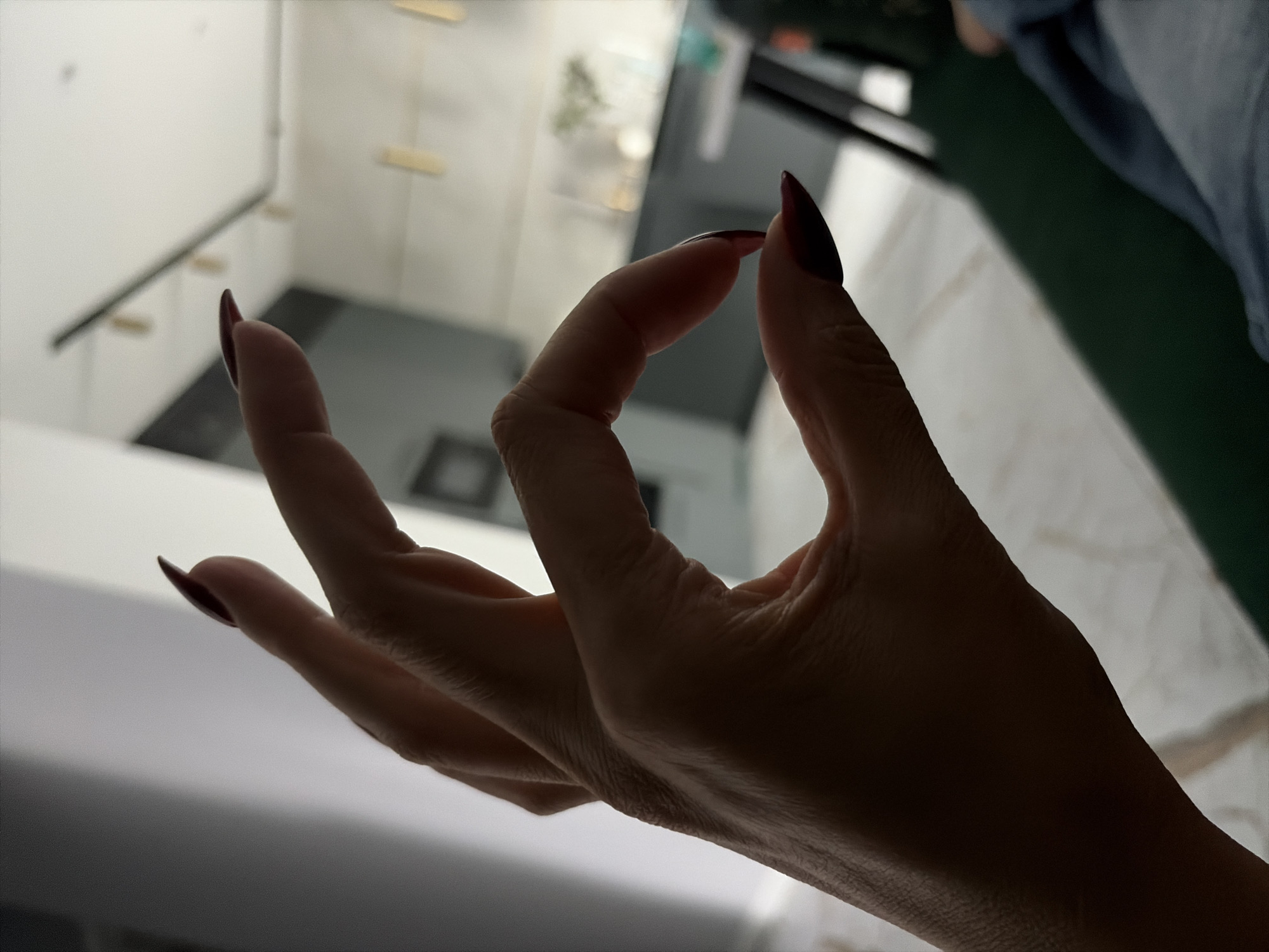 A close-up, silhouetted shot shows a woman's right hand forming an "okay" gesture. Her fingers, painted with dark red nail polish, are curved to create a circle. The background is blurred, revealing a modern interior with what appears to be a kitchen area, track lighting on the ceiling, and furniture. The lighting in the image is dim and comes from the left, casting the hand in shadow and highlighting the edges of her fingers. The location is not specified.