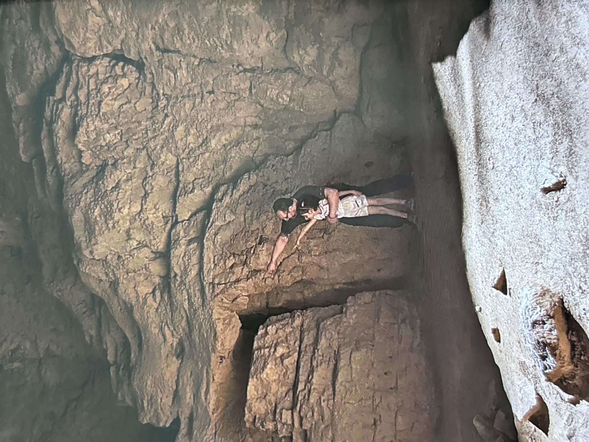 The image portrays two individuals, an adult male and a child or young person, exploring a rugged cave or rock formation. The adult, wearing a dark t-shirt and light-colored shorts, is positioned in the foreground, leaning forward with his right arm extended downwards into a dark, narrow opening or crevice in the rocky floor or ledge. He is looking into this opening with focus. The child, dressed in a light-colored top and patterned shorts, stands directly behind the adult, appearing to be held or supported by him, and is also looking towards the dark crevice.
The setting is characterized by massive, textured rock walls and surfaces, predominantly in shades of brown and gray, displaying numerous cracks, layers, and rough details. The lighting is dim and uneven, casting deep shadows that highlight the geological features and create a sense of depth within the enclosed space. To the right of the frame, a distinct, lighter-colored rock formation, appearing almost white or sandy, stands in contrast to the darker main rock face. This lighter formation has several small, triangular indentations or holes.
The activity depicted is one of cautious exploration and discovery within a natural, subterranean environment. No specific time of day or weather conditions are discernible due to the indoor, cave-like setting. There is no visible text in the image.