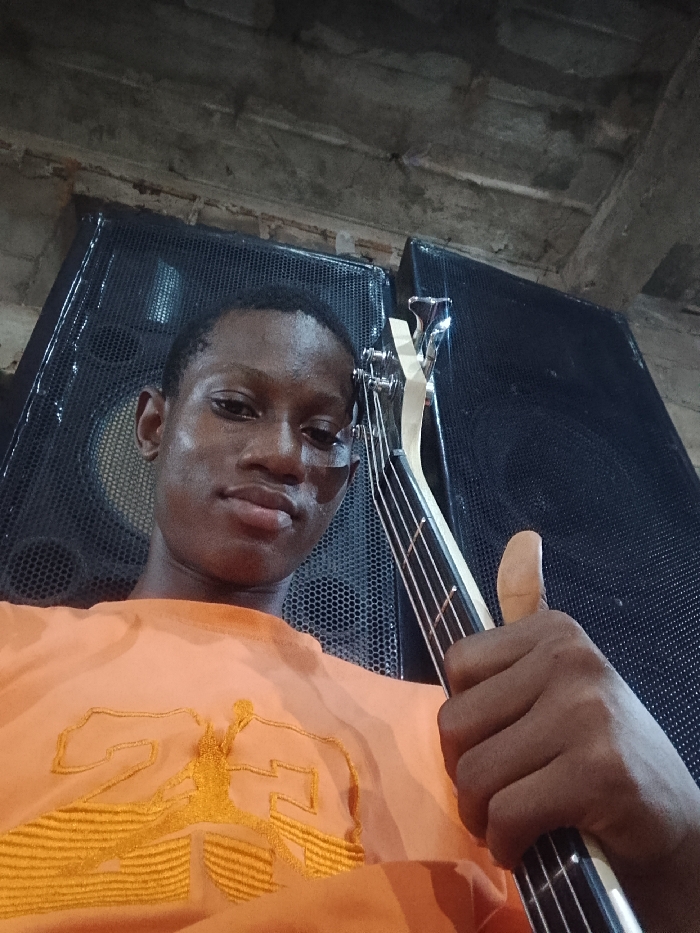 A selfie features a young man holding a four-string bass guitar, giving a thumbs up. He's wearing an orange shirt with an embroidered design, possibly a logo. Behind him are two large, dark speaker cabinets. The background suggests an indoor setting with a textured or unfinished ceiling. The lighting appears somewhat dim and uneven, creating shadows on the subjects and the surroundings.
