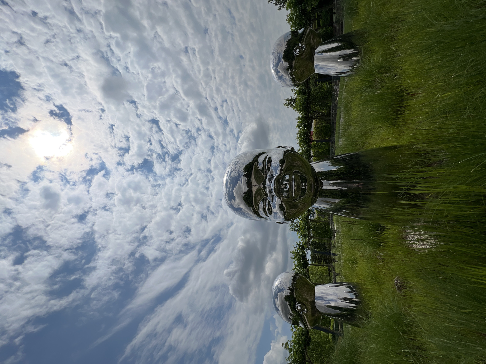 The image shows three large, shiny, metallic sculptures of baby heads, positioned in a grassy field. The central sculpture is larger and faces directly towards the viewer, with a smiling expression. The other two sculptures are smaller and positioned on either side of the central one, facing slightly away. They are made of polished metal that reflects the sky and the surrounding environment. The sky above is filled with white, fluffy clouds, and the sun is visible through a gap in the clouds, casting a bright light. In the background, there is a row of trees and a dark fence. The grass in the foreground is tall and green. This scene is located in the city of Krasnodar, Russia.
