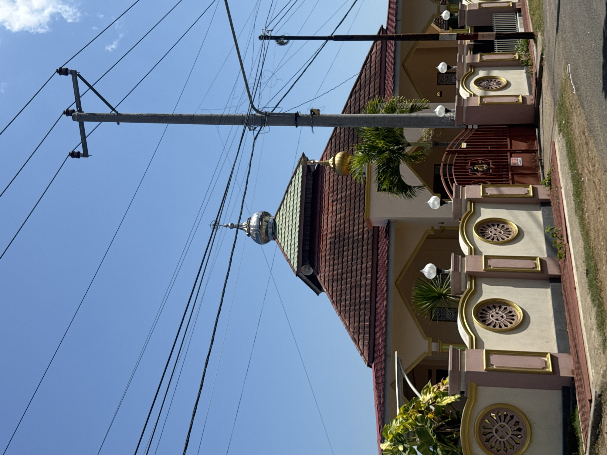 In the city of Kertasari, Indonesia, an Islamic mosque stands beneath a clear blue sky, crisscrossed by a network of power lines and supported by a concrete utility pole. The mosque features a distinctive brown tiled roof and a green dome topped with a silver spire. A smaller golden dome is visible on the right side of the building. A decorative pink and gold fence surrounds the property, punctuated by circular ornamental windows. A large, maroon metal gate is set within the fence. Lush green foliage, including a palm tree, grows around the mosque and along the fence line. The scene is observed from across a paved road.