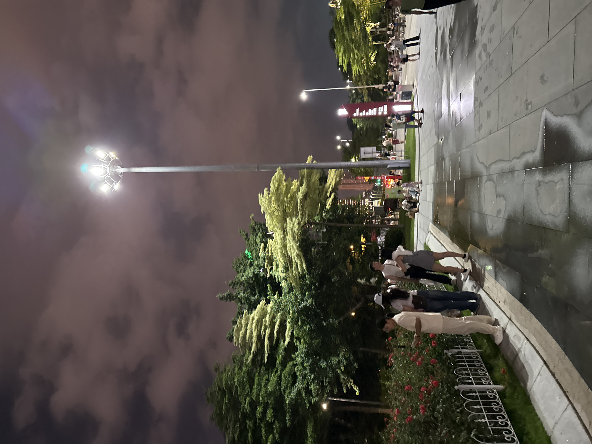 The image captures an outdoor scene at night, possibly in Tianhe District, China. The sky is dark with scattered, illuminated clouds, suggesting a cloudy night. A tall lamppost with multiple lights stands prominently in the foreground, casting a bright light on the surroundings. Several people are visible in the image, appearing to be walking or gathered on a paved area. Some are standing in groups, while others are walking individually or in pairs.
The ground is paved with large, rectangular tiles, and there are patches of wetness, indicating recent rain or a damp surface. Lush green trees and some flowering plants, possibly roses, are present, adding a touch of nature to the urban setting. The lighting is artificial, coming from the lamppost and other unseen sources, creating a somewhat dramatic ambiance.
The people are dressed casually, and their postures suggest a relaxed atmosphere. Some appear to be looking at their phones, while others are engaged in conversation. The overall mood seems calm and peaceful, typical of a leisurely evening in a public space. There is no discernible text visible in the image. The presence of the lamppost and the paved walkways suggest this might be a park, plaza, or a public walkway in a city.
