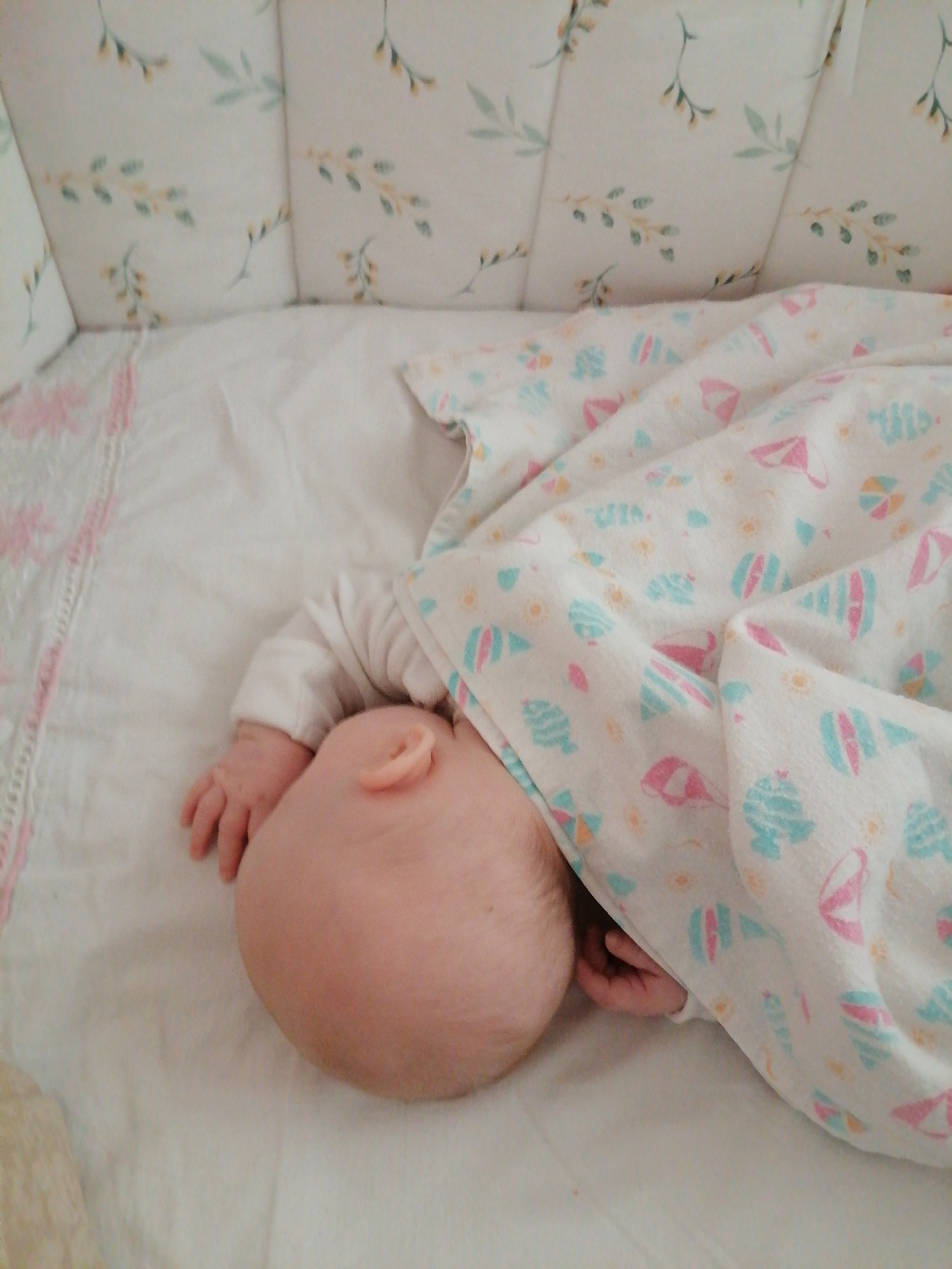 The image shows a baby sleeping in a crib. The baby is lying on a white sheet with a pink patterned border. A blanket with a pattern of beach-themed images, in pink and light blue, covers part of the baby. The baby is wearing a white long-sleeved shirt. Only the back of the baby's head, one ear, and part of one arm are visible. The crib has patterned bumpers with a design of light-colored foliage. The location is unknown.