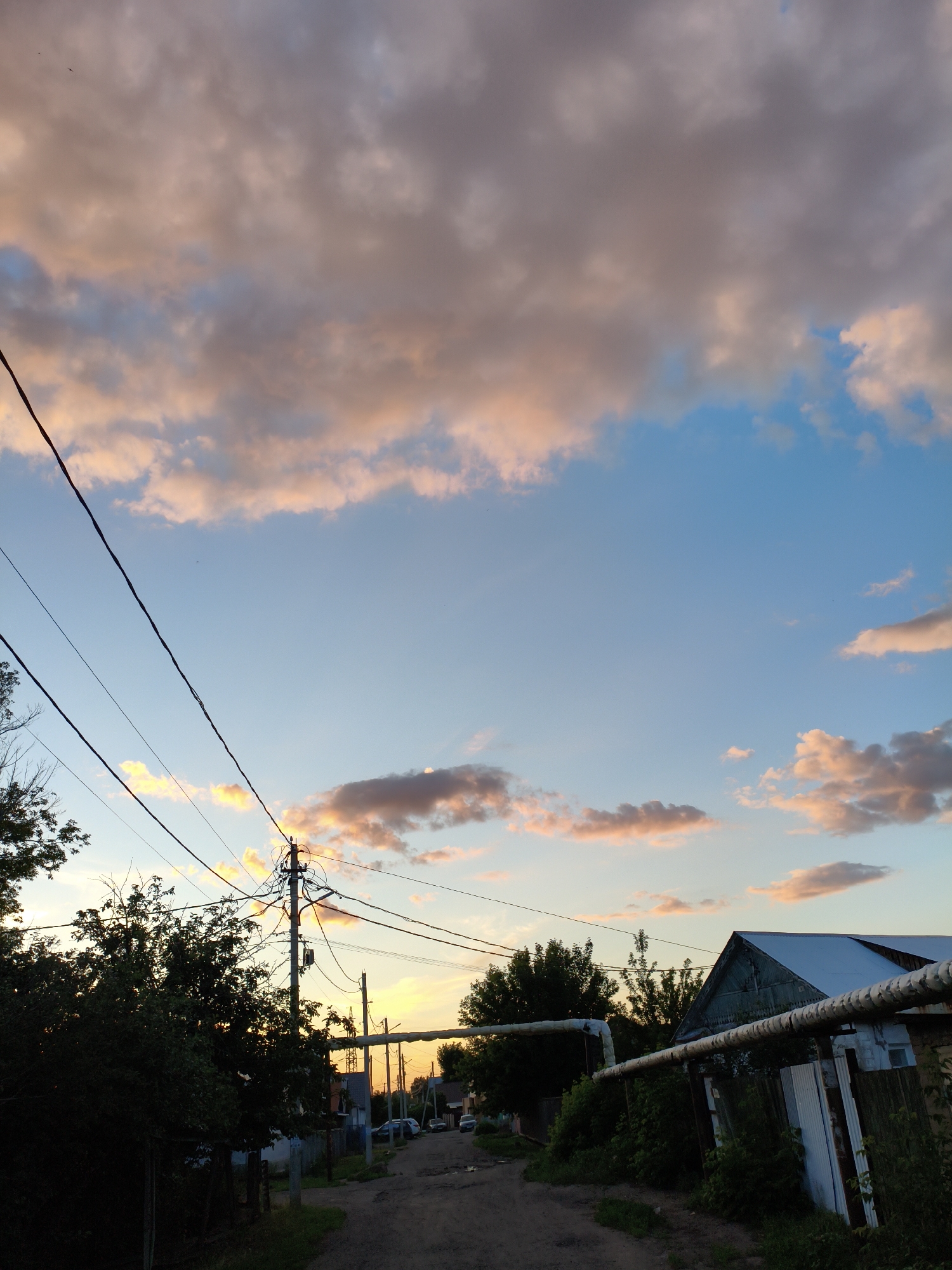 The image depicts a rural or suburban street scene in Orenburg, Russia, during what appears to be sunset. The sky is a prominent feature, filled with a mix of blue sky and clouds tinged with pink and orange hues, indicating the time of day. Several black utility wires crisscross the frame, strung between utility poles, with one pole being particularly complexly wired.
On the left side of the image, a dense cluster of dark green trees is visible, obscuring any potential structures behind them. A dirt or gravel road runs through the center of the image, leading into the distance. Several vehicles, including cars and possibly a minivan, are parked or driving along this road.
On the right side of the road, a light-colored building with a corrugated metal roof is partially visible. Adjacent to this building is a metal fence and a large, insulated pipe running horizontally across the foreground. The pipe appears to be part of an above-ground utility system. The overall impression is a modest, somewhat unkempt environment, typical of many residential areas outside of major city centers. There are no people clearly visible in the image, and no overt actions are taking place. The scene is static and observational.