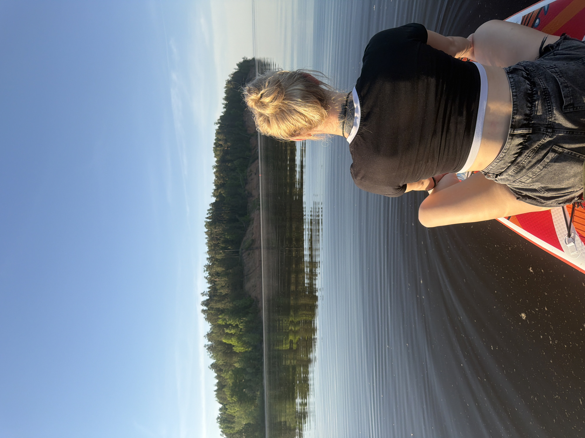 A person sits on a stand-up paddleboard on calm water with a forested shoreline in the background under a clear blue sky. The individual has blonde hair tied in a bun, and is wearing a black top and gray denim shorts. The water reflects the forest and sky. The location is Noviy, Russia.