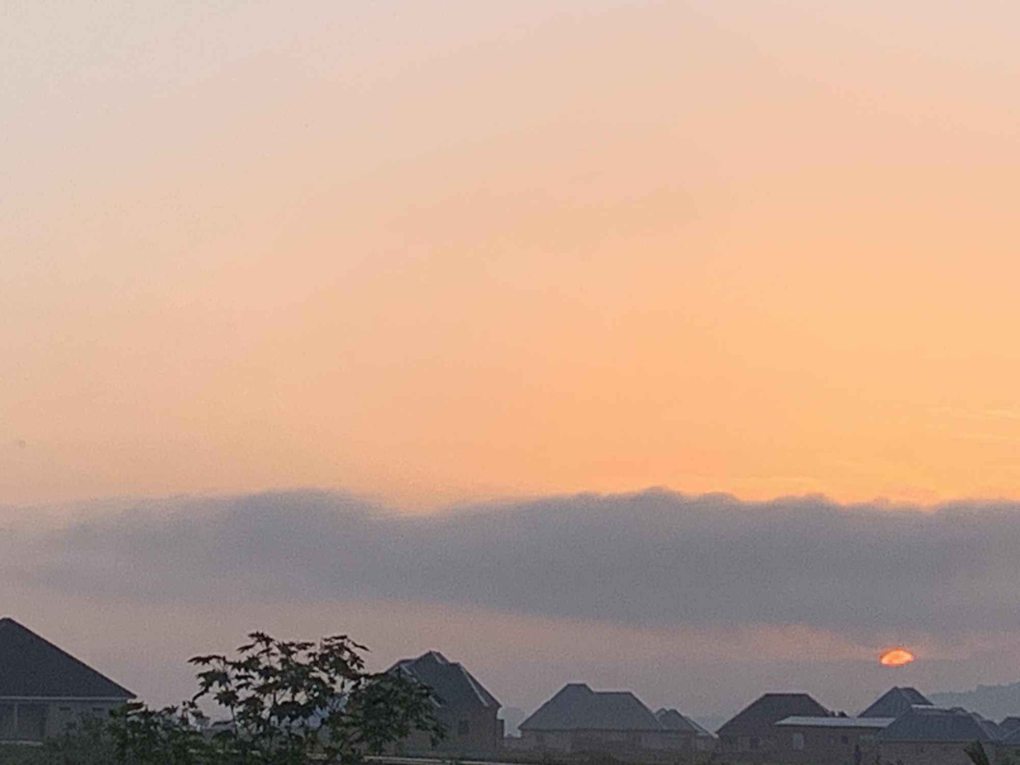 The image captures a sunrise over a residential area in Kuru Karama, Nigeria. The sky is a gradient of soft peach and pale orange, with a band of low-lying gray clouds stretching horizontally across the middle of the frame. Partially obscured by these clouds, the sun appears as a vibrant orange disc just above the horizon. Silhouetted against the sky are the dark, angular roofs of several houses, indicating a built-up area. In the foreground, the silhouette of a leafy tree is visible on the left, adding a touch of natural greenery to the scene. The overall atmosphere is serene and quiet, typical of the early morning hours.