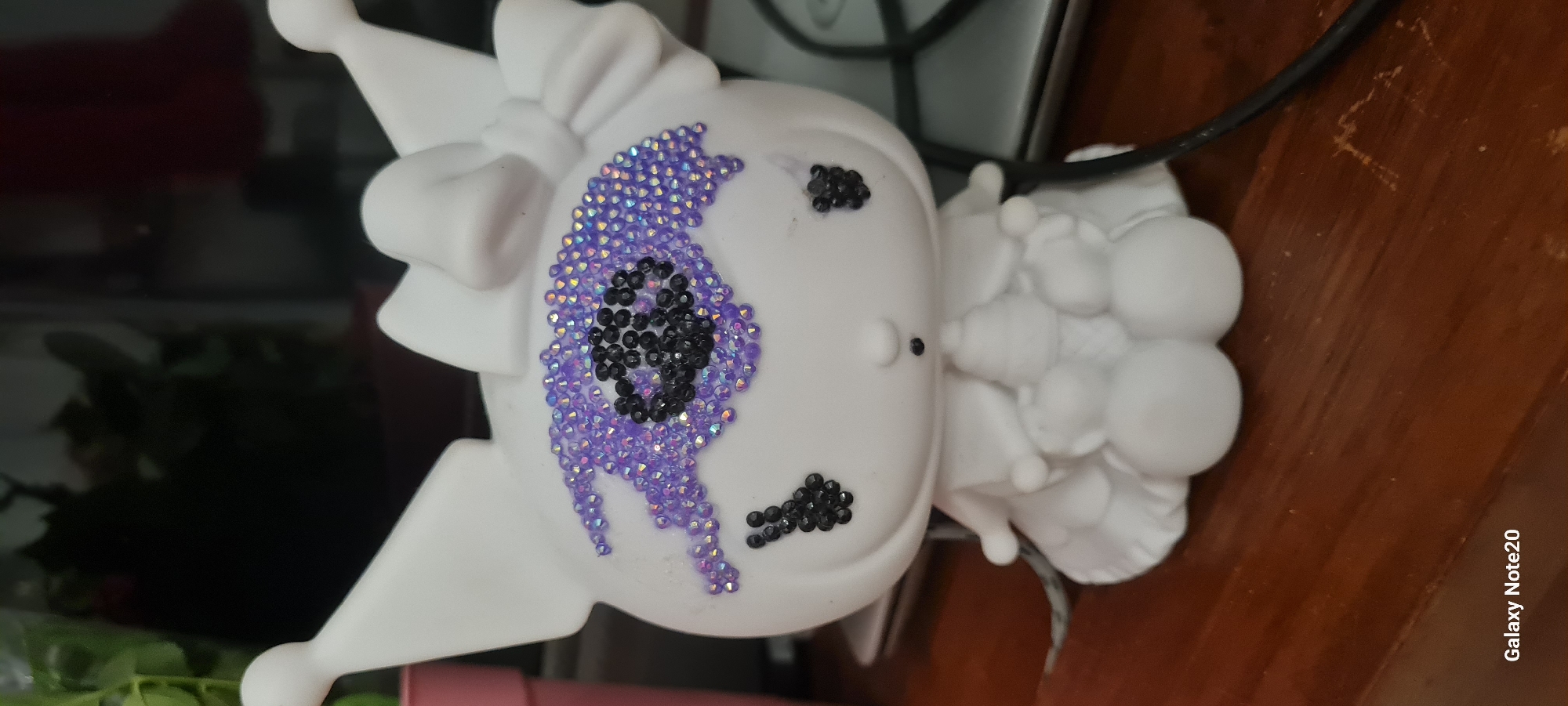 A close-up, eye-level, portrait shot shows a white ceramic figurine of a cat-like character on a wooden surface. The figurine has large ears, a bow on its head, and a dress-like lower body. Its face is decorated with black beads forming the pupils of its eyes and a small nose. Around its eyes, it has purple rhinestones arranged in an eyeshadow pattern. The figurine is holding what appears to be ice cream cones in its arms. The background is blurred, showing hints of greenery and possibly a dark reflection. A portion of a black cable and a silver surface are visible to the right of the figurine. The bottom left corner of the image displays the text "Galaxy Note20". The scene is likely indoors, possibly in a home environment. There is no information to suggest the location is in the city of Holon, Israel.